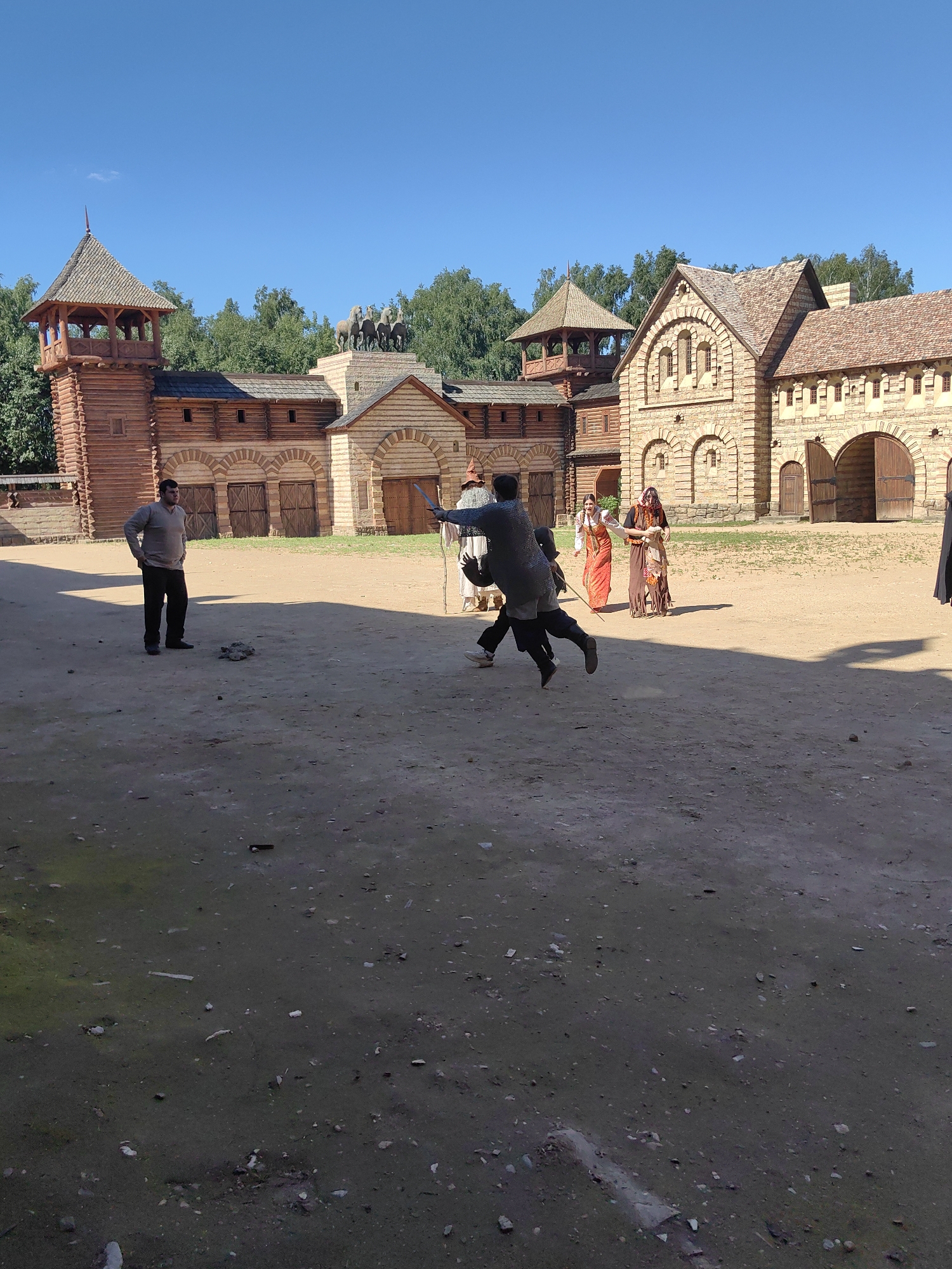 A mid-shot captured outdoors on a sunny day shows a historical reenactment or film set. The background features a fortified wooden structure with towers and stone buildings, reminiscent of a medieval settlement. In the foreground, several individuals are present. Two men are engaged in a sword fight, one in chainmail is mid-lunge. Another man in a light-colored shirt and dark pants stands watching the fight. Two women in traditional historical clothing are also present, observing the scene. The ground is dirt and sand, with scattered debris. The sky is clear and blue. The location is identified as Терехово-2 Снт, Russia.