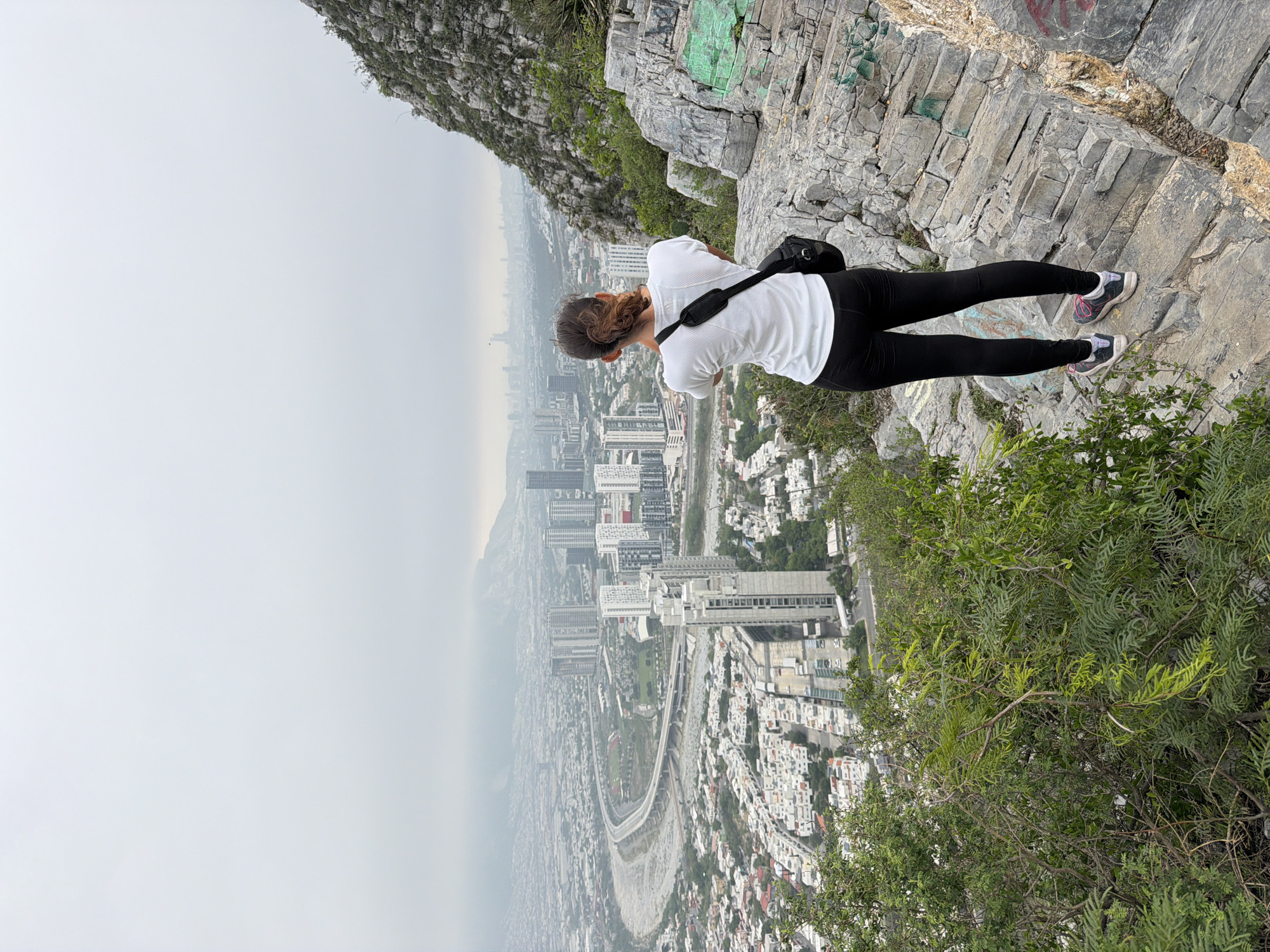 A medium shot shows a woman wearing a white t-shirt and black leggings standing on a rocky cliff edge, leaning forward to look out over the city of Santa Catarina, Mexico. The woman is facing away from the camera, and a black strap is visible across her back. The landscape below is hazy, with buildings and roads stretching into the distance. The cliff edge is composed of gray rocks and sparse vegetation.