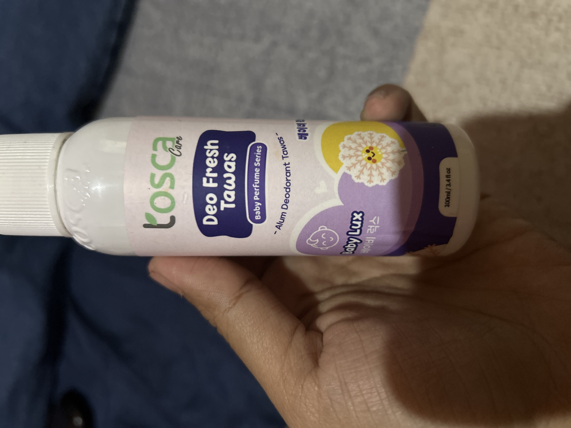 A hand is holding a white plastic spray bottle of Tosca Care Deo Fresh Tawas, a baby perfume series deodorant. The bottle has purple and pink labels with text and illustrations. The background appears to be a dark blue fabric and a textured light-colored surface. The overall scene is indoors, likely in a residential setting. There is no specific information to indicate the location is Depok, Indonesia.