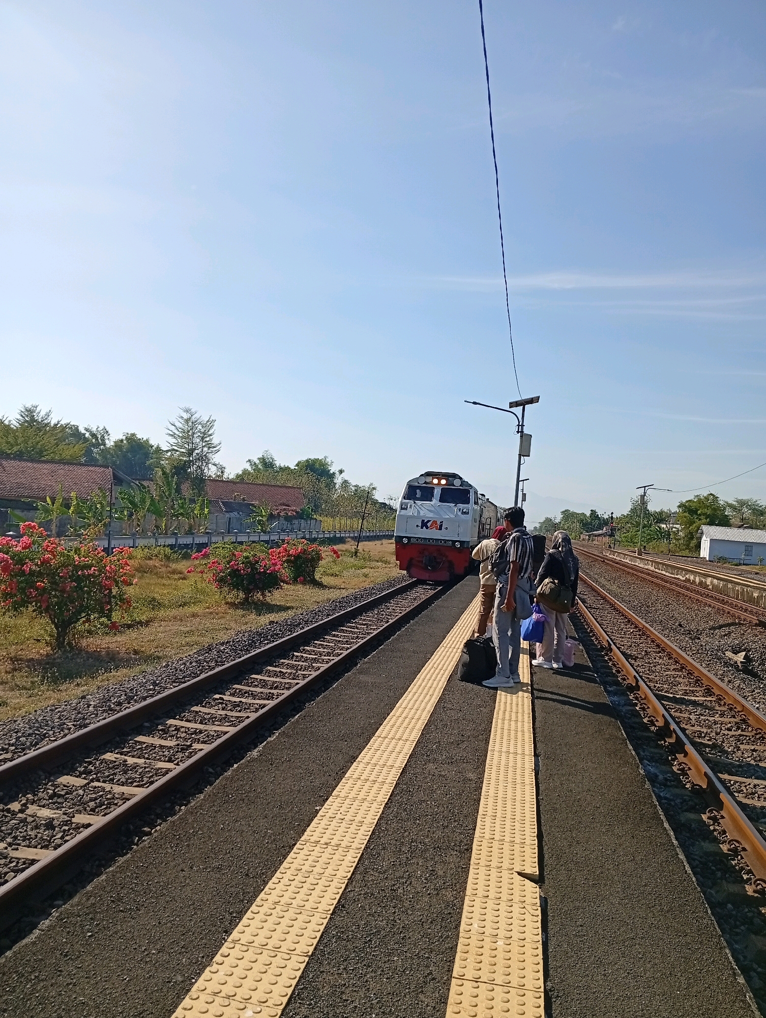 A train is stopped at a station platform in Tebon, Indonesia, under a clear blue sky. The train is white and red with "KAI" logo visible. A group of four people, three women and one man, stand on the platform with luggage, appearing to be waiting. The platform is paved with asphalt and has a tactile paving strip. There are two sets of train tracks, with gravel ballast. In the background, there are houses with red tiled roofs, trees, and bushes with pink flowers. A solar-powered street light stands on the platform. The overall scene depicts a quiet moment at a rural Indonesian train station.