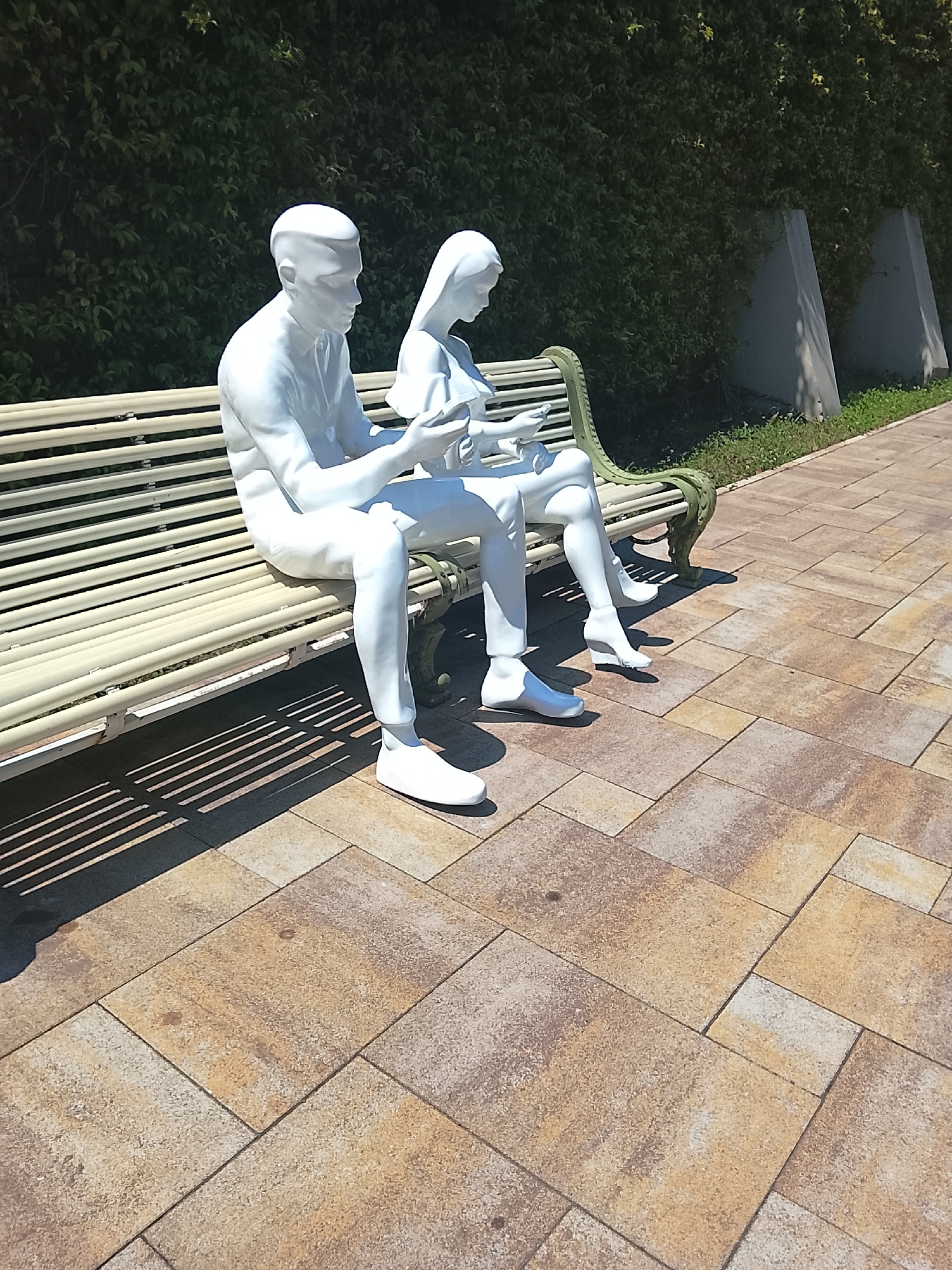 This image depicts two white, life-sized statues of a man and a woman seated on a light-colored park bench. The man is on the left, and the woman is on the right. Both statues are engaged in looking at or holding objects in their hands, suggesting they are reading or interacting with mobile devices. The bench has a green, ornate metal frame.
The statues are positioned on a paved walkway made of rectangular, multi-colored stone tiles in shades of brown, tan, and gray. To the left of the bench, the ground is partially covered by the bench's shadow, which forms distinct linear patterns. Behind the bench, there is a dense green hedge acting as a backdrop. Two triangular, light-colored concrete structures are visible behind the hedge, to the right of the statues. The environment suggests an outdoor public space, possibly a park or a promenade.
There is no information available to confirm or deny the location in the city of Oryol-Izumrud, Russia.