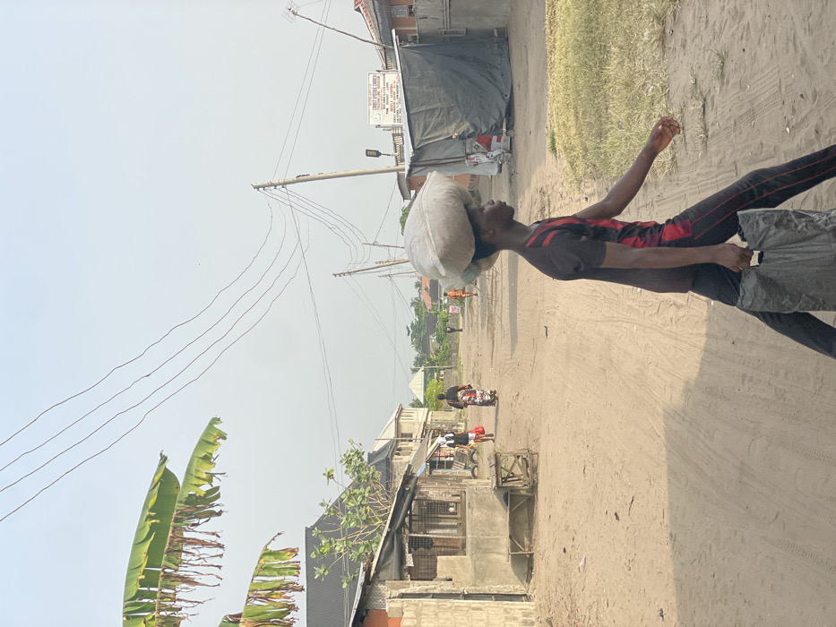 This image depicts a daytime scene on an unpaved, sandy road in Mosafejaw, Nigeria. The central figure is a young male, seen in profile, walking from right to left across the frame. He carries a large, light-colored sack or bundle balanced on his head and a dark, possibly plastic, shopping bag in his right hand. He wears a dark t-shirt with red accents and dark trousers with red piping.
In the mid-ground, other individuals are visible on the road: a person in a white top walking left with a red bag, and behind them, a person in a patterned dress walking away from the camera. Further down the road, a person on a motorcycle travels away from the viewer, and several distant figures are also present.
The environment includes single-story and multi-story buildings lining both sides of the road. On the left, a concrete block structure adjoins a wooden stall with a corrugated roof, featuring a wooden bench in front. On the right, utility poles with multiple overhead wires run along the roadside. A white sign for "CHRIST APOSTOLIC CHURCH" with "MOSHAFEJA BRANCH" is legible in the distance on the right, above a structure covered by a dark tarp. Overhanging banana leaves are visible in the upper left, and the sky is a hazy, light blue or grey. Long shadows across the road suggest morning or late afternoon light.