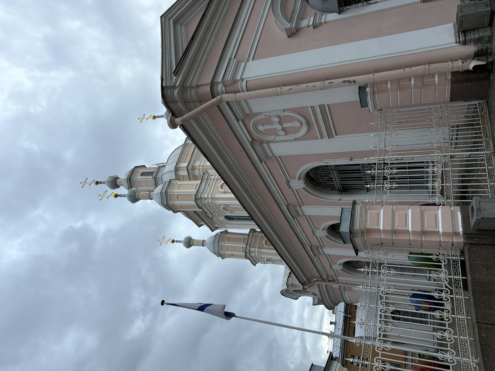 The image shows the Smolny Cathedral in Saint Petersburg, Russia, viewed from a low angle, with a cloudy sky overhead. The pink facade of the cathedral features white architectural details, arched windows with iron grates, and several onion domes topped with golden crosses. A white flag with blue stripes, likely a naval or ensign flag, flies on a tall flagpole to the left of the cathedral. In the foreground, a decorative white metal fence encloses the area. Behind the fence, two large, colorful spherical objects, one blue and one green, are partially visible, suggesting some kind of temporary installation or event. The ground is paved with cobblestones.