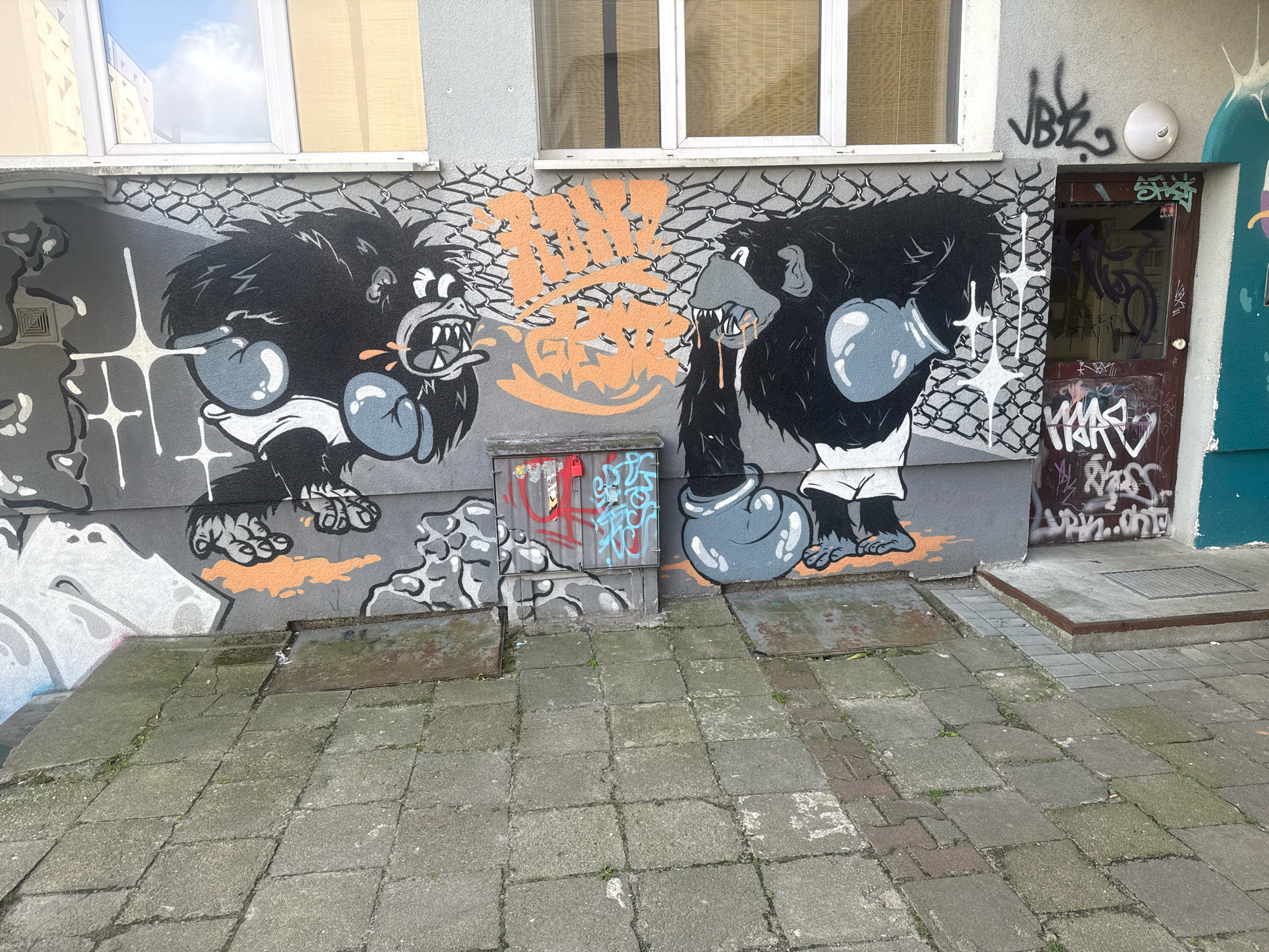 The image displays graffiti on the exterior wall of a building in Gorzów Wielkopolski, Poland. Two cartoonish gorillas, depicted in a boxing stance with large boxing gloves, are the central subjects of the graffiti. The gorilla on the left is spitting with its mouth open, while the gorilla on the right appears to be drooling. Between the two gorillas, there is orange graffiti with a lightning bolt design. To the right of the graffiti, there is a brown door with more graffiti on it, and a white circular object mounted on the wall above the door. The foreground consists of a paved walkway made of square concrete slabs, some of which are cracked and covered in moss. The wall itself is gray, with a chain-link fence element incorporated into the design between the two gorillas. Above the graffiti, there are windows of the building. The overall scene depicts an urban environment with street art.
