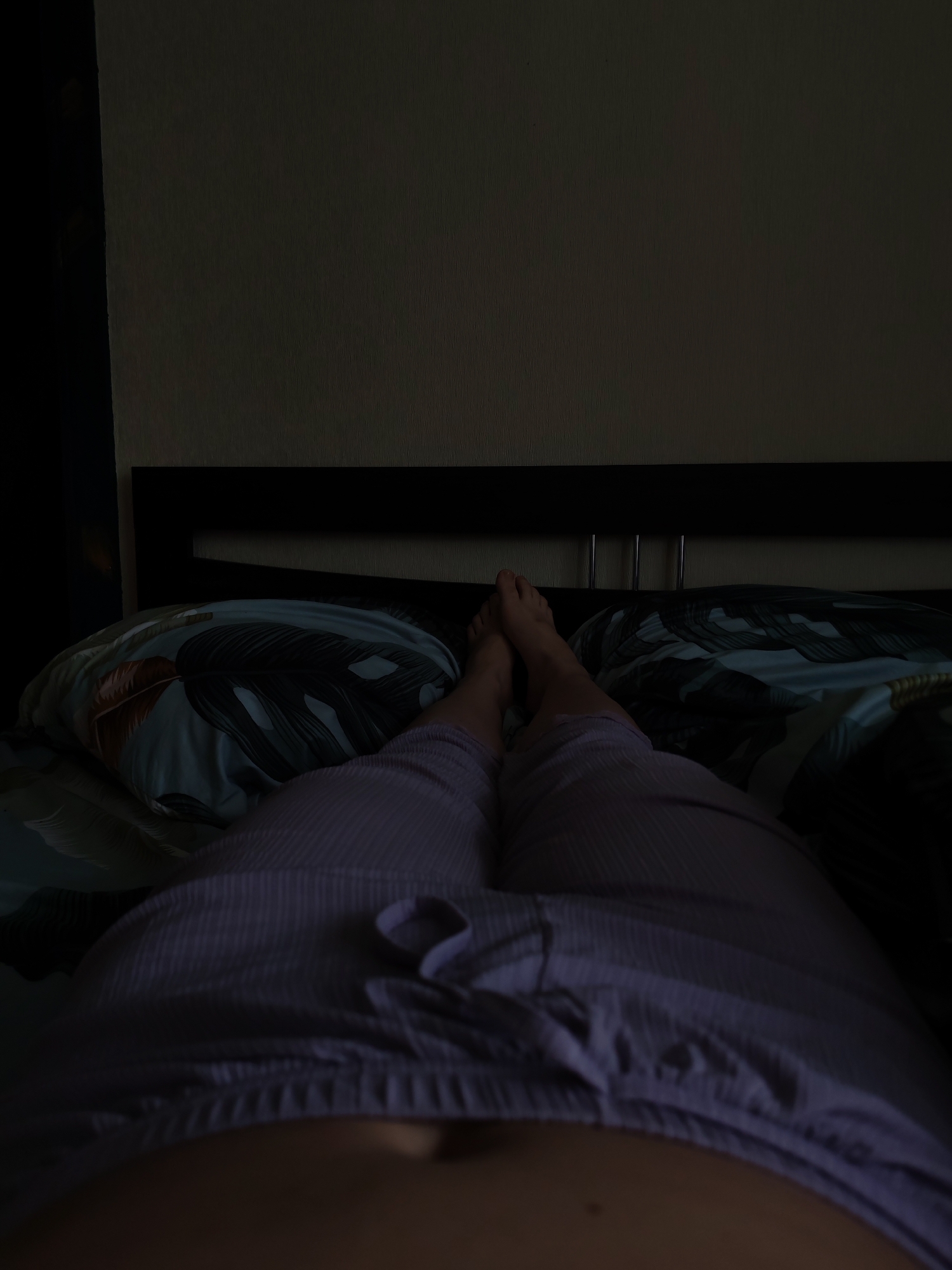The image shows a person lying in bed in a dimly lit room. The person is wearing lilac-colored pajama pants with a drawstring at the waist. Their feet are visible near the headboard of the bed, which is dark brown or black. The bedding includes pillows with a tropical leaf pattern. The room has a wall painted in a light neutral color. The exact location within Izhevsk, Russia, cannot be determined from the image.