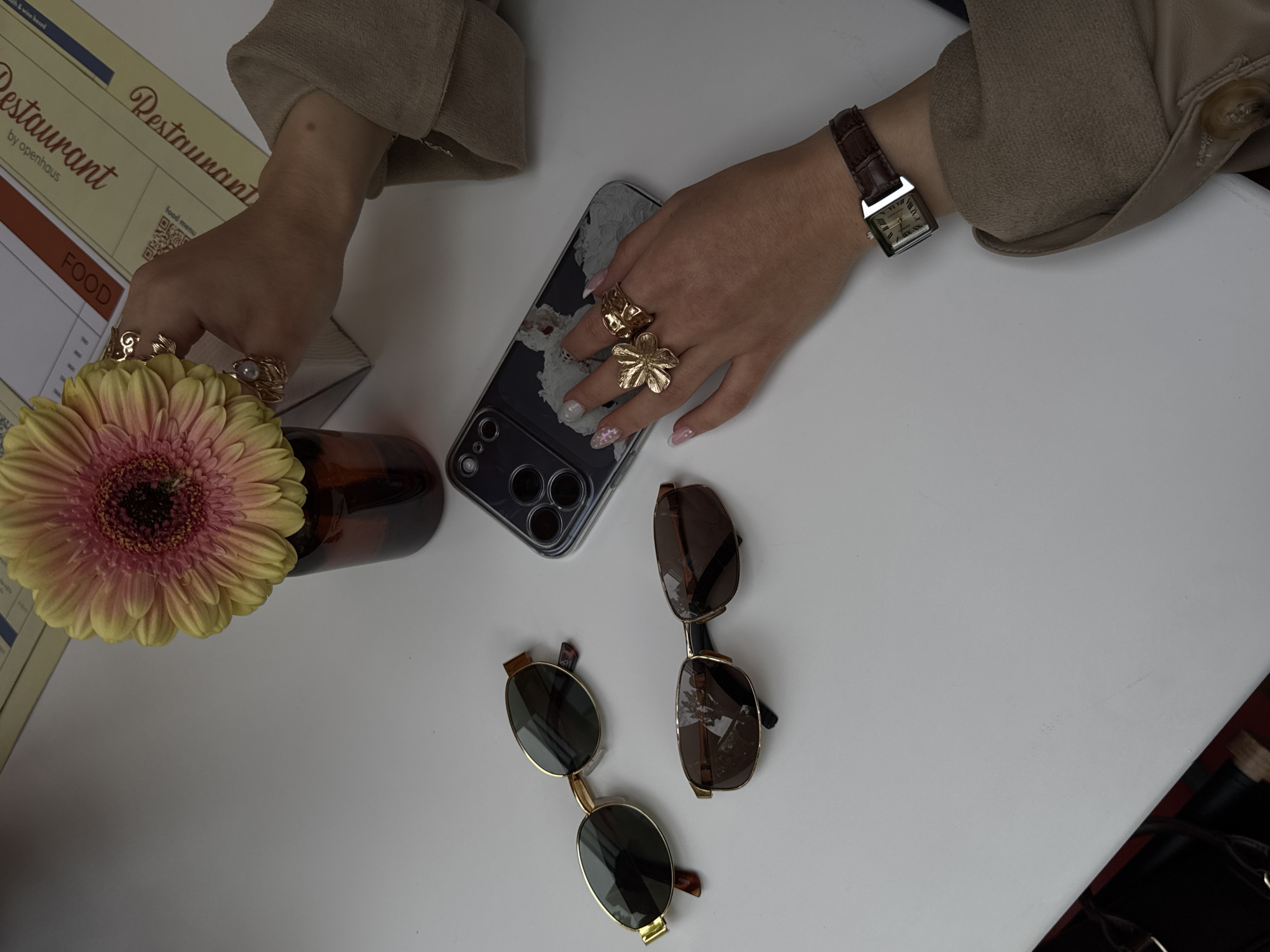 The image captures a tabletop scene in a restaurant, presumably located in Yerevan, Armenia, judging by the "Restaurant by openhaus" branded menu visible in the upper left. A woman's hands are the central focus. The left hand holds a gerbera daisy flower in a brown glass vase. The right hand rests near a cell phone with a clear case featuring a cloudy pattern. The woman wears a brown leather-strapped rectangular watch on her right wrist and multiple gold rings on both hands. Two pairs of sunglasses lie next to the phone. The background is a plain white surface.
