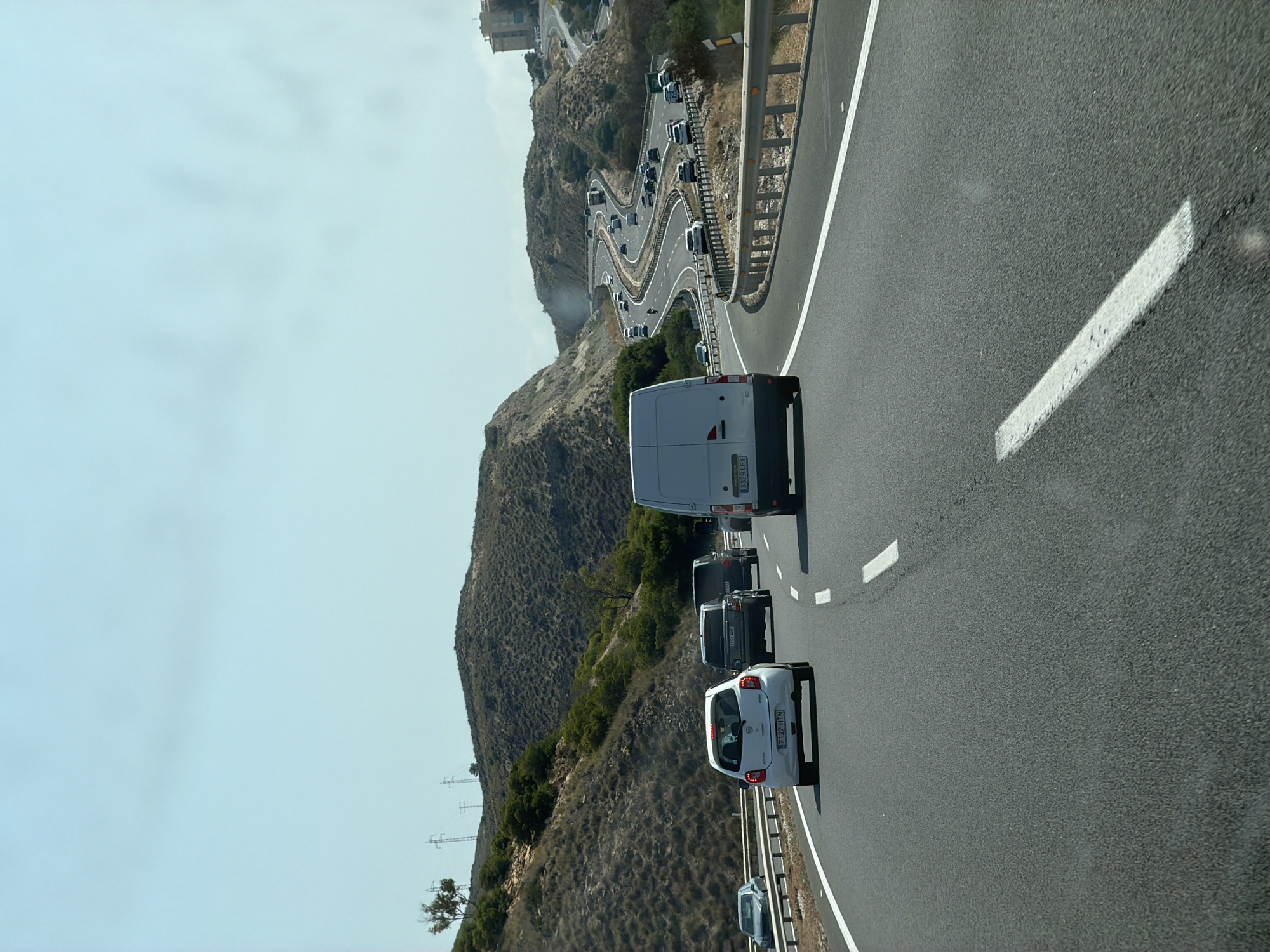 The image shows a view from a vehicle traveling on a multi-lane highway. The highway curves to the right, with rolling, dry hills on either side, dotted with sparse vegetation. Several vehicles are visible on the highway, including a white hatchback in the foreground, a dark SUV and a dark van further ahead, and a white van on the right side of the frame. The sky is clear and pale blue. In the distance, a multi-story building is visible on a hilltop to the right. The scene appears to be in a warm, dry climate.