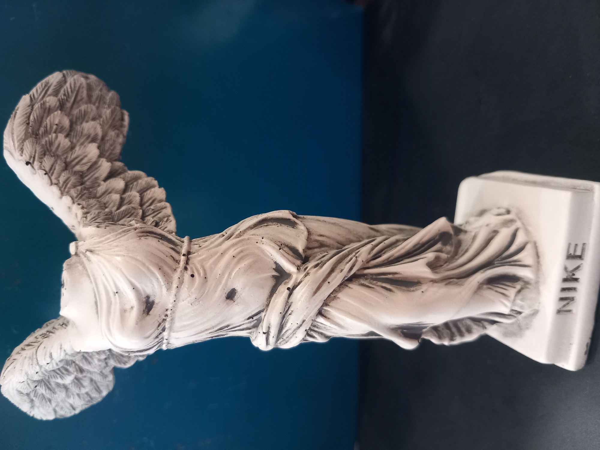 A headless, white statue of Nike, the Greek goddess of victory, stands on a dark surface against a deep blue background. The statue is depicted in motion, with her wings spread and her draped garment flowing. The base of the statue has the word "NIKE" inscribed on it. The statue is positioned slightly off-center to the left of the frame, with the right side of the statue extending towards the edge of the image. The lighting casts subtle shadows and highlights on the statue, emphasizing its form and texture. The overall scene is one of artistic representation and historical homage.