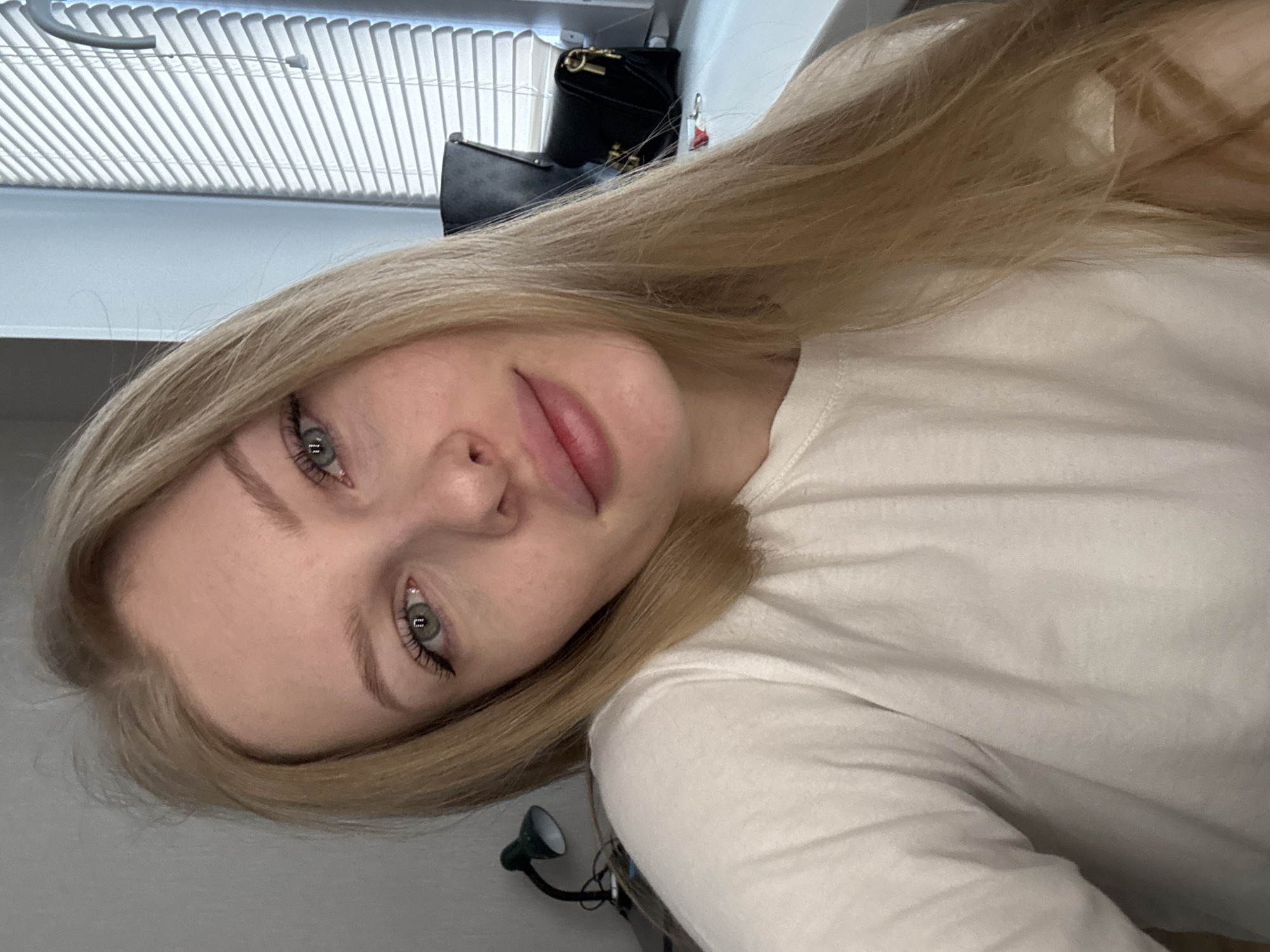 A close-up, angled portrait of a fair-skinned woman with blonde hair and green eyes. She is wearing a white long-sleeved shirt. The background includes a window with blinds, and a black bag is visible. A small lamp is on the wall. There is no indication of the city of Perm, Russia.
