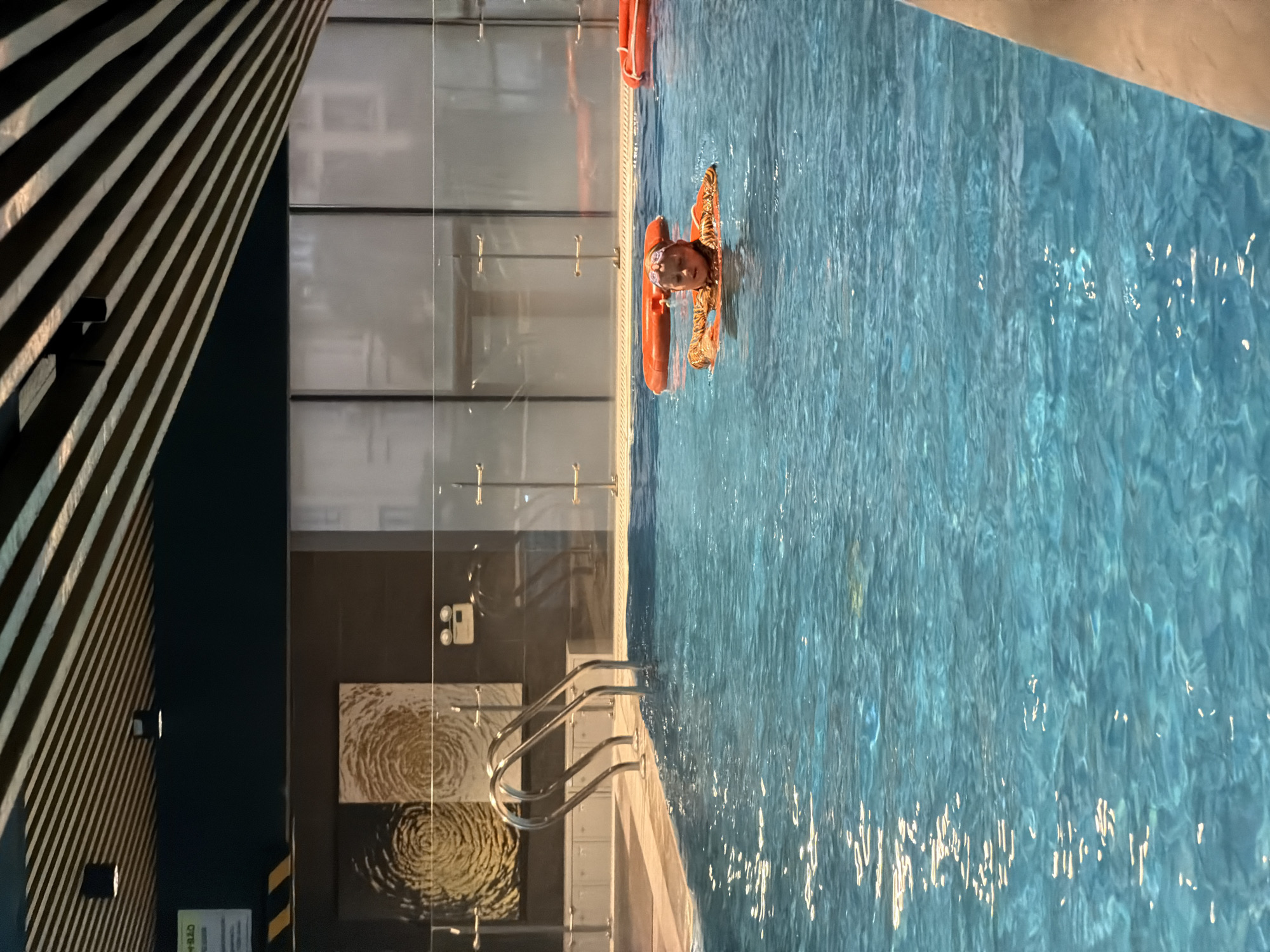 A young girl with blonde hair, wearing goggles and an orange swim vest with a tiger pattern, is swimming in a bright blue indoor pool. She is positioned in the center of the frame, looking towards the camera. The pool water is clear and reflects sunlight. In the background, there are several orange inflatable rings floating on the surface of the water. The pool area appears to be modern, with a dark ceiling featuring striped panels and subtle lighting. There are glass barriers around the pool, and two abstract artworks with spiral designs hang on a dark wall to the left. To the right of the artwork, a set of silver pool stairs with a curved handle is visible at the edge of the water. The overall environment suggests a public or private indoor swimming facility, possibly a hotel or a recreational center, but the specific location in Hà Nội, Vietnam cannot be determined from the image.