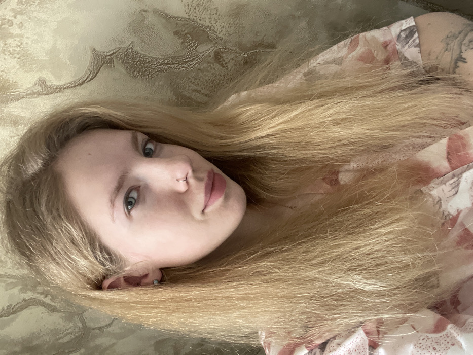 A fair-skinned person with long, straight blonde hair lies on a textured surface. Their head is slightly tilted, showcasing light blue eyes, a nose piercing, and soft pink lips. The person is wearing a floral-patterned top. A glimpse of a tattoo is visible on their upper arm. The background has a subtle, embossed design.