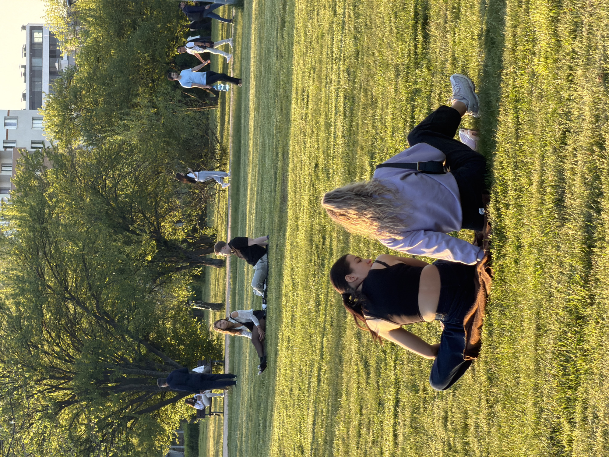 The image captures a vibrant, sunlit scene in an urban park, likely during the late afternoon or early evening "golden hour," given the warm, directional light and long shadows cast across the green grass. The setting features a wide expanse of lawn, interspersed with numerous deciduous trees, and multi-story buildings are visible in the background, suggesting an urban environment like Sofia, Bulgaria.
In the foreground, two young women are seated casually on the grass. One, with long, wavy blonde hair, wears a light purple long-sleeved top, dark trousers, and white sneakers. She is looking towards the second woman, who has dark hair tied back in a ponytail and is dressed in a black tank top and dark pants, with a brown garment draped over her lap. They appear relaxed, observing their surroundings.
Further back, a larger group of people is scattered across the park. Many individuals appear to be engaged in outdoor physical activity or a communal gathering. One man, wearing a light blue t-shirt and dark pants, stands with slightly outstretched arms, a large clear water bottle visible near his feet. Another man in a black t-shirt and light pants is seen bending over. Other figures are standing, walking, or sitting on benches in the middle ground and background, blending into the park's tranquil atmosphere. The lush trees form a natural barrier, separating the active area from the distant buildings. The overall impression is one of leisure and activity in a peaceful public green space. There is no visible text in the image.