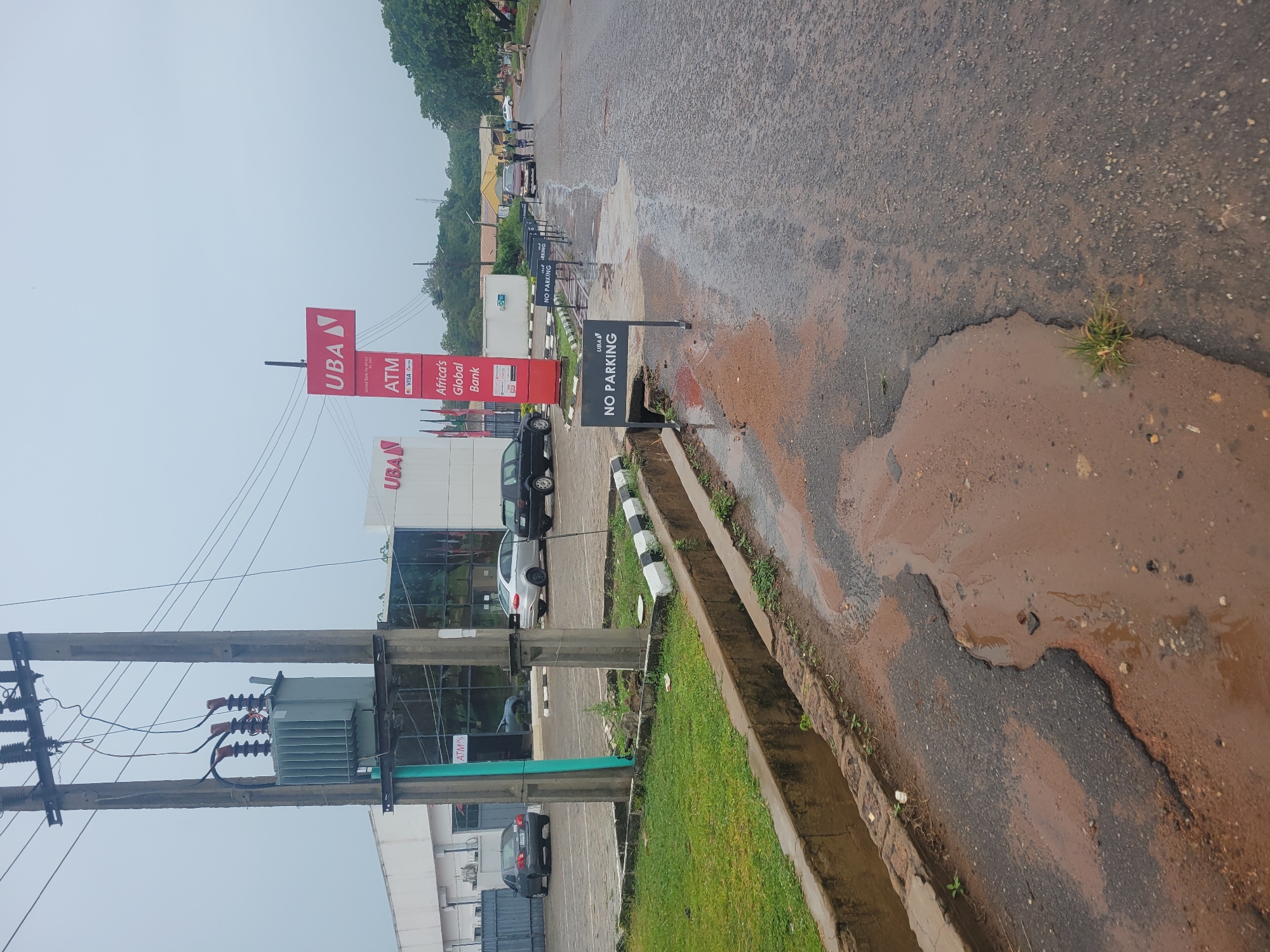 The image shows a street scene outside a UBA bank branch. A large electrical transformer is visible on a utility pole. Several cars are parked in the bank's lot. A line of "NO PARKING" signs is placed along the side of the road. The road itself appears wet and has puddles of water, suggesting recent rain. Some pedestrians are walking down the street in the distance. The UBA building has a modern facade with large windows. The sky is overcast.