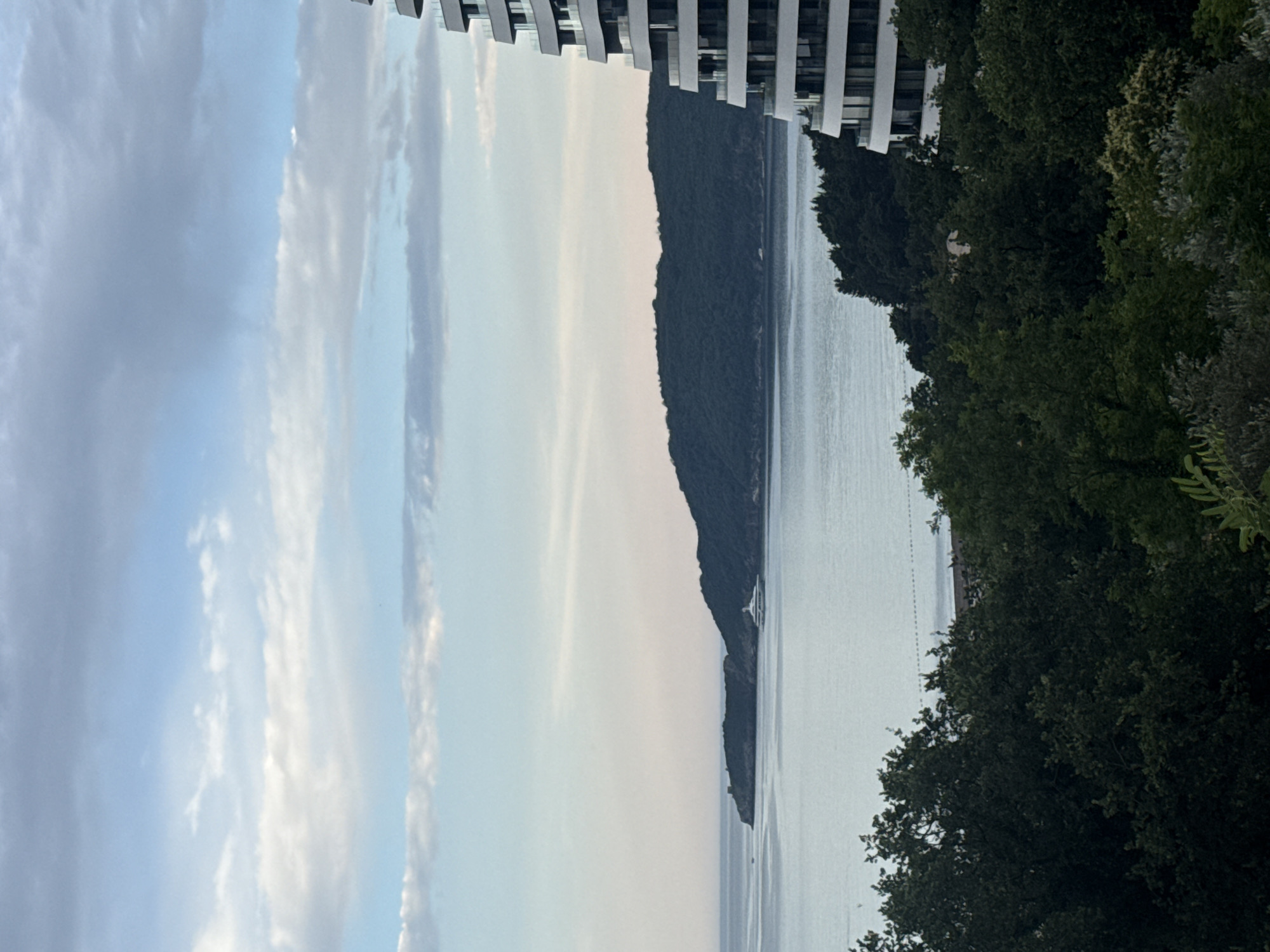 This image depicts a scenic view of a body of water, likely a bay or sea, adjacent to a lush, tree-covered landmass. The water is calm and reflects the soft, pastel hues of the sky, suggesting it is either early morning or late evening. Wispy clouds drift across the sky, adding texture to the serene atmosphere.
In the foreground, on the right side of the image, a dense line of dark green trees creates a natural frame. Their foliage appears healthy and vibrant. A modern building with a white facade and horizontal dark elements is visible at the top left of the image, suggesting proximity to a developed area.
A prominent feature in the middle ground is a steep, dark green hillside that slopes down towards the water, creating a dramatic natural outline. On this hillside, near the water's edge, a small white structure, possibly a pavilion or a boat, is visible. The water stretches out towards the horizon, where it meets a hazy sky. In the distance, along the shoreline, a faint line of what appears to be a beach or a breakwater can be discerned.
The overall mood of the image is peaceful and tranquil, with a sense of natural beauty enhanced by the presence of the modern architecture. The lighting suggests a soft, diffused light, characteristic of dawn or dusk. No text is visible in the image. The location context of Boreti, Montenegro, aligns with the depiction of a coastal landscape with hills and water.