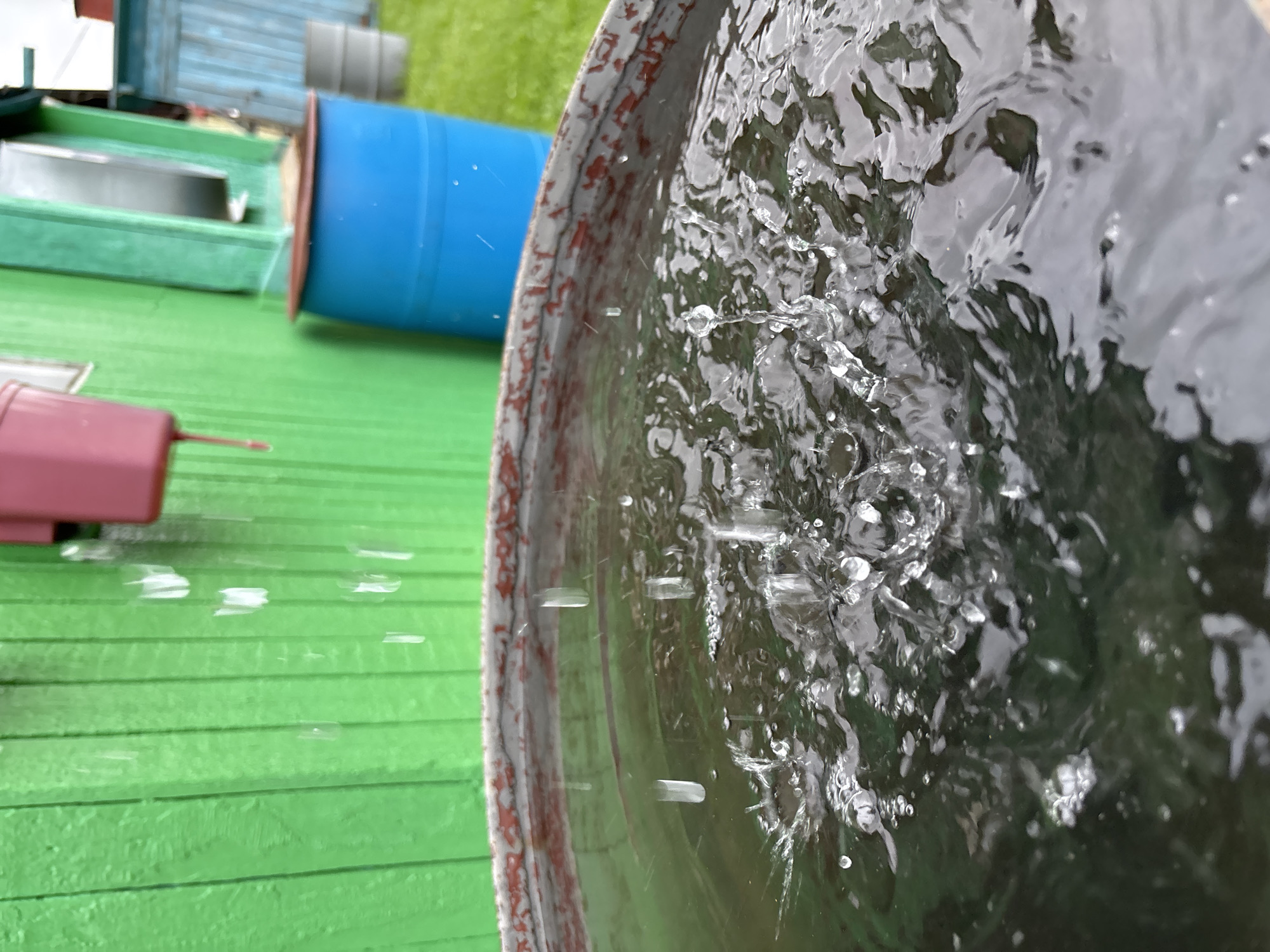 In Gonja, Russia, water is seen dripping from a pink downspout onto the surface of a large, rustic metal barrel filled with dark water. The barrel is positioned in front of a vibrant green wooden wall. Behind the barrel, a bright blue plastic barrel stands, and further in the background, a gray barrel is visible, alongside a blue structure. The ground appears to be covered in green grass. The focus is on the water droplets creating ripples and splashes as they impact the surface of the water in the foreground barrel, suggesting an active rainfall or water collection scenario.