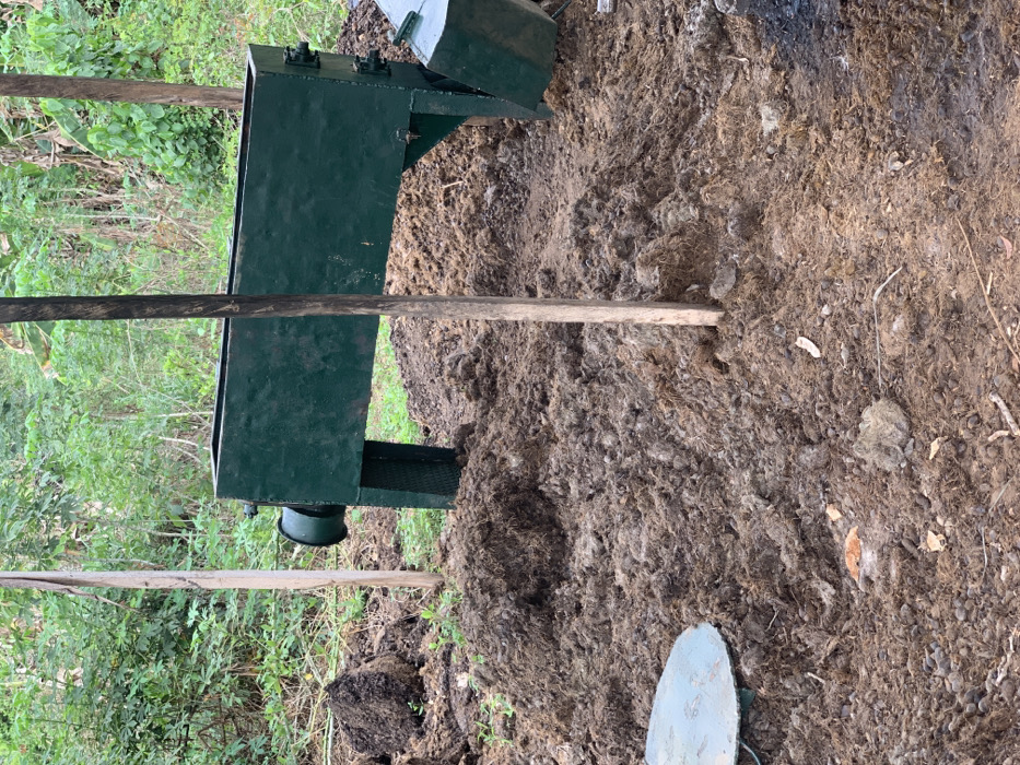 The image depicts a green metal structure, likely a composting or manure processing device, situated in a densely vegetated area. The structure rests on a bed of brown organic material, likely a mixture of manure and other compostable waste. A wooden pole is positioned diagonally across the frame, partially obscured by the vegetation. The structure is located in a rural area of Egbeda, Nigeria, at coordinates 7.119 latitude, 5.362 longitude.