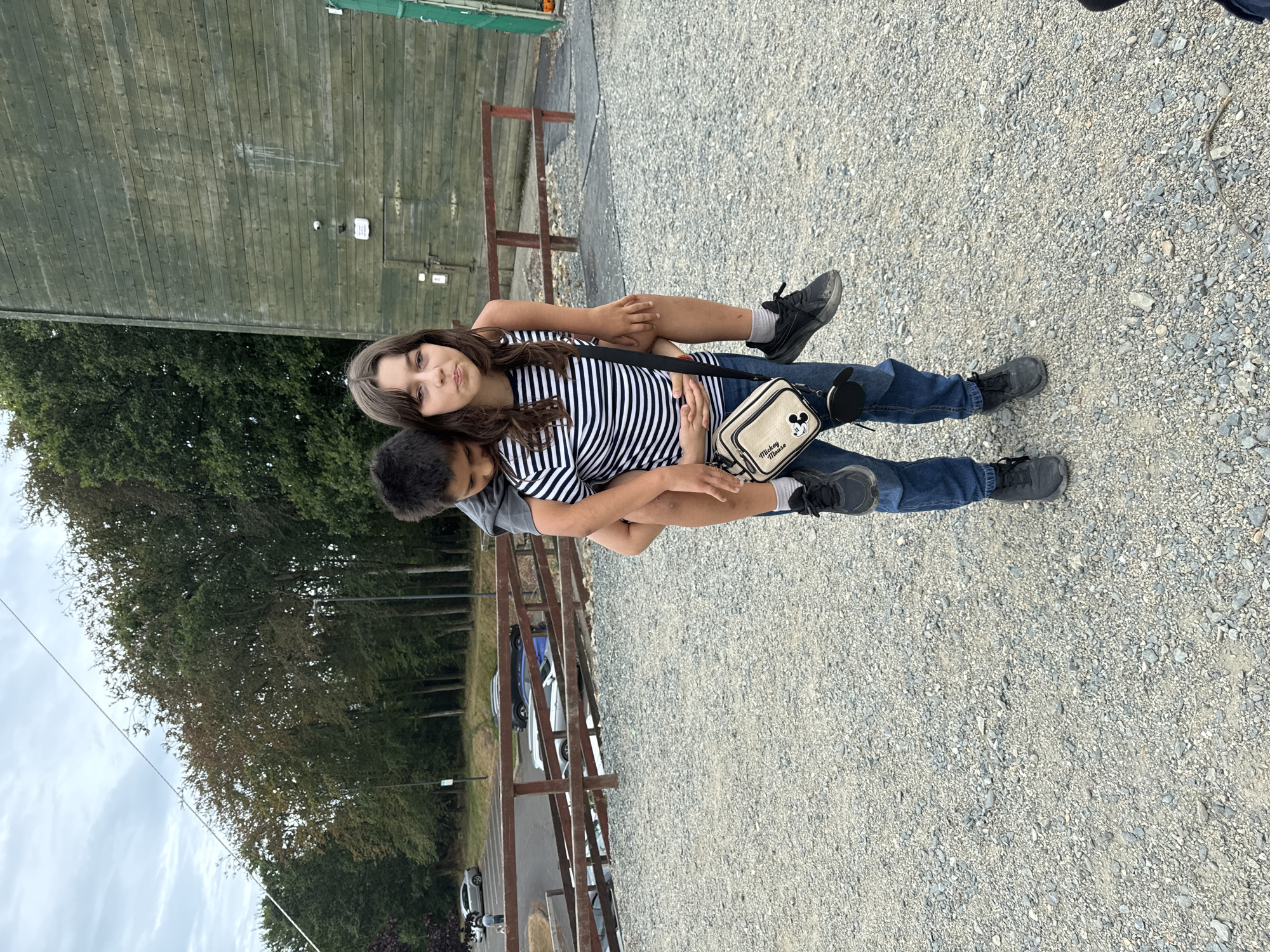 The image shows two young people outdoors on a gravel path. A boy is giving a girl a piggyback ride. The girl is wearing a black and white striped t-shirt, blue jeans, and black sneakers. She has a small beige bag with a Mickey Mouse design hanging across her body. The boy is wearing a grey t-shirt and blue jeans. They appear to be in a park or recreational area, with a wooden fence and trees in the background. To the left of the fence, there is a large wooden structure. In the distance, several cars are parked. The sky is cloudy, suggesting it might be an overcast day. The overall mood seems relaxed and playful. The image is oriented vertically, with the subjects positioned centrally.