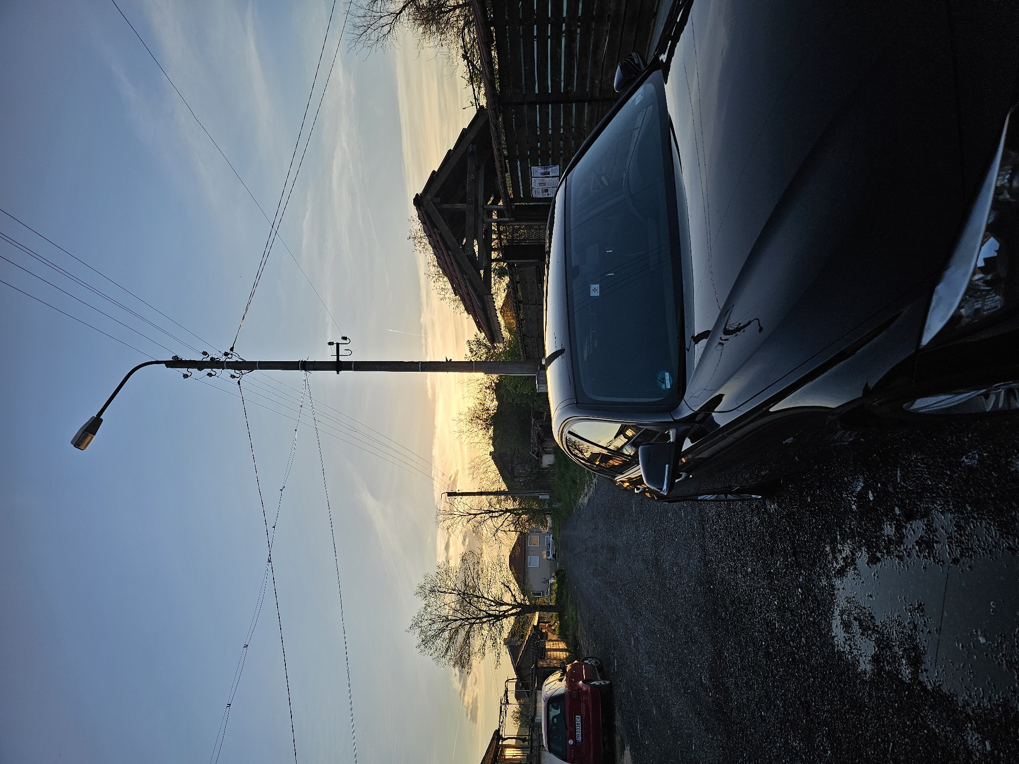The image shows an outdoor scene in Krasnovo, Bulgaria. A black car is parked to the right. To the left of the car is a gravel road and a red car further down the road. A street light and utility pole are visible along the left side of the frame, stretching up towards the sky. In the background, there are trees, a house, and a wooden structure. The sky displays a gradient of colors, suggesting either sunset or sunrise. The road appears wet, indicating recent rain.