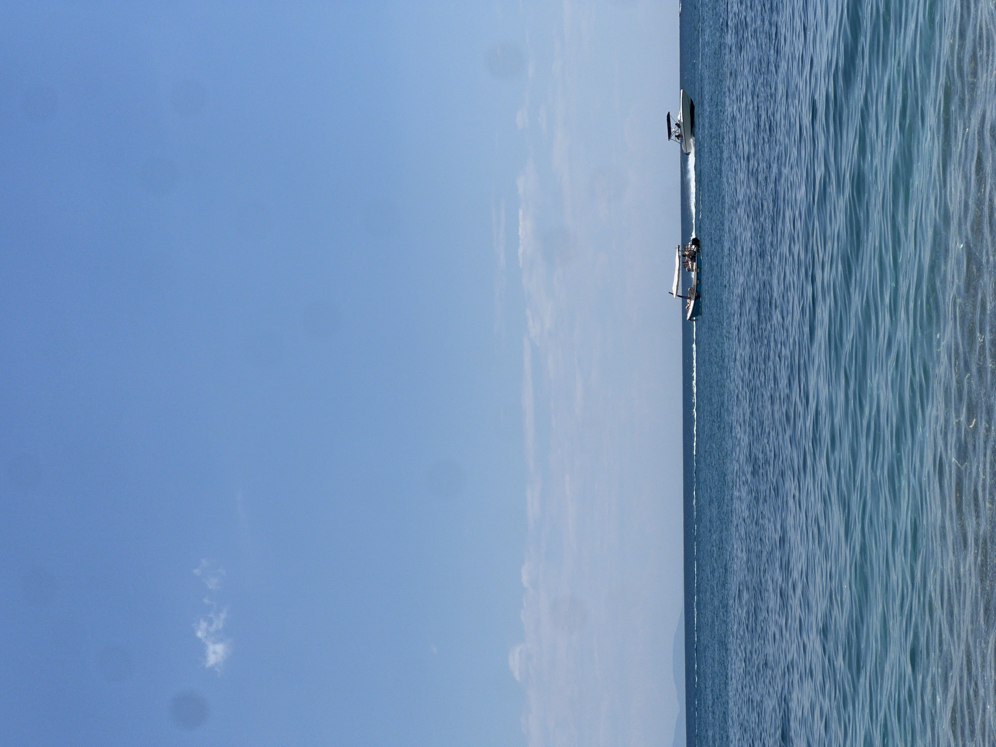 The image depicts a seascape, with a clear blue sky occupying the majority of the left side and the ocean on the right. Two boats are visible on the water. One is a small sailboat with what appears to be people aboard, while the other is a larger motorboat traveling at a higher speed. The photograph appears to have been taken at the location of Prijevor, Montenegro, with the coastline and open water extending out into the distance.