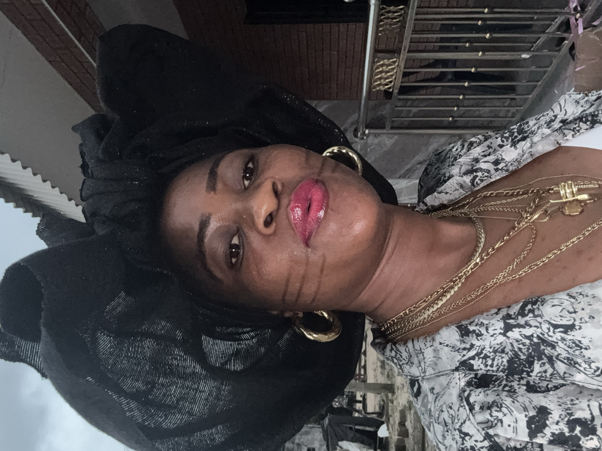 In this medium shot, a Black woman, viewed from the chest up, poses outdoors. She is wearing a large black fascinator hat adorned with textured fabric, multiple gold necklaces with a cross pendant, a patterned jacket with black and white designs, and large gold hoop earrings. The woman has dark skin, full lips painted with glossy pink lipstick, and her eyes are looking towards the camera. She is standing outside a building with a red brick facade and a metal railing in the background. To the left of the frame, there is a glimpse of an outdoor area with what appears to be tarps and structures. The lighting suggests it is daytime, possibly overcast. The location is identified as Ajara, Nigeria.