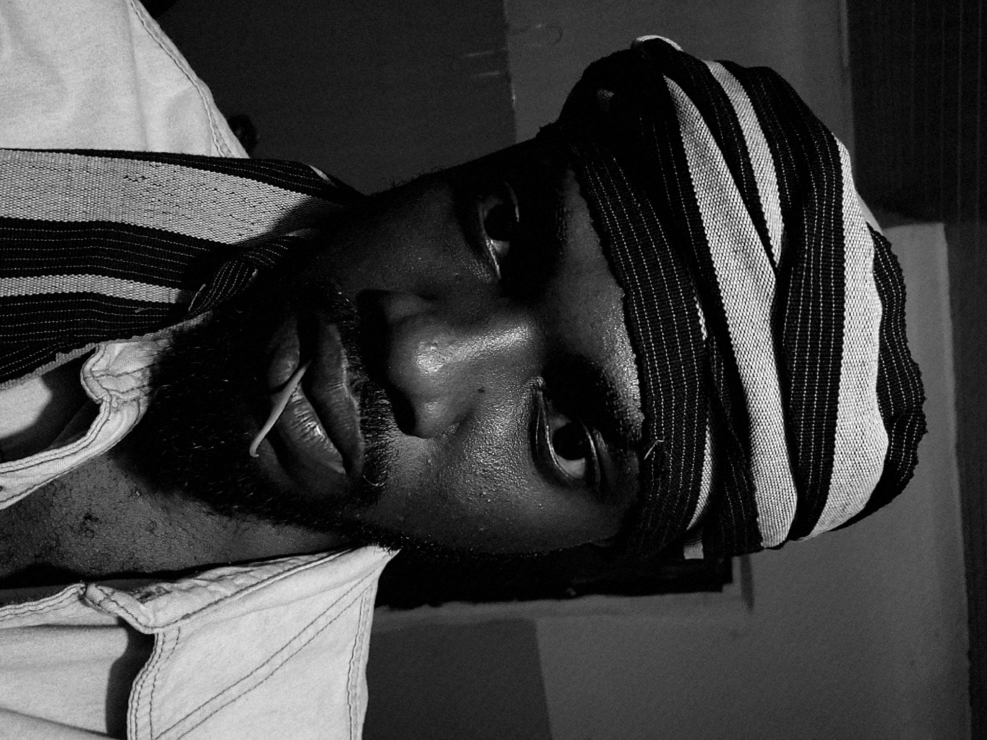 A close-up, black and white portrait shows a dark-skinned man wearing a striped headwrap. He has a beard and mustache. The man is wearing a collared shirt. His face is slightly tilted. The location in the city of Jos, Nigeria, cannot be definitively determined based solely on this image. The environment is dimly lit and somewhat indistinct.