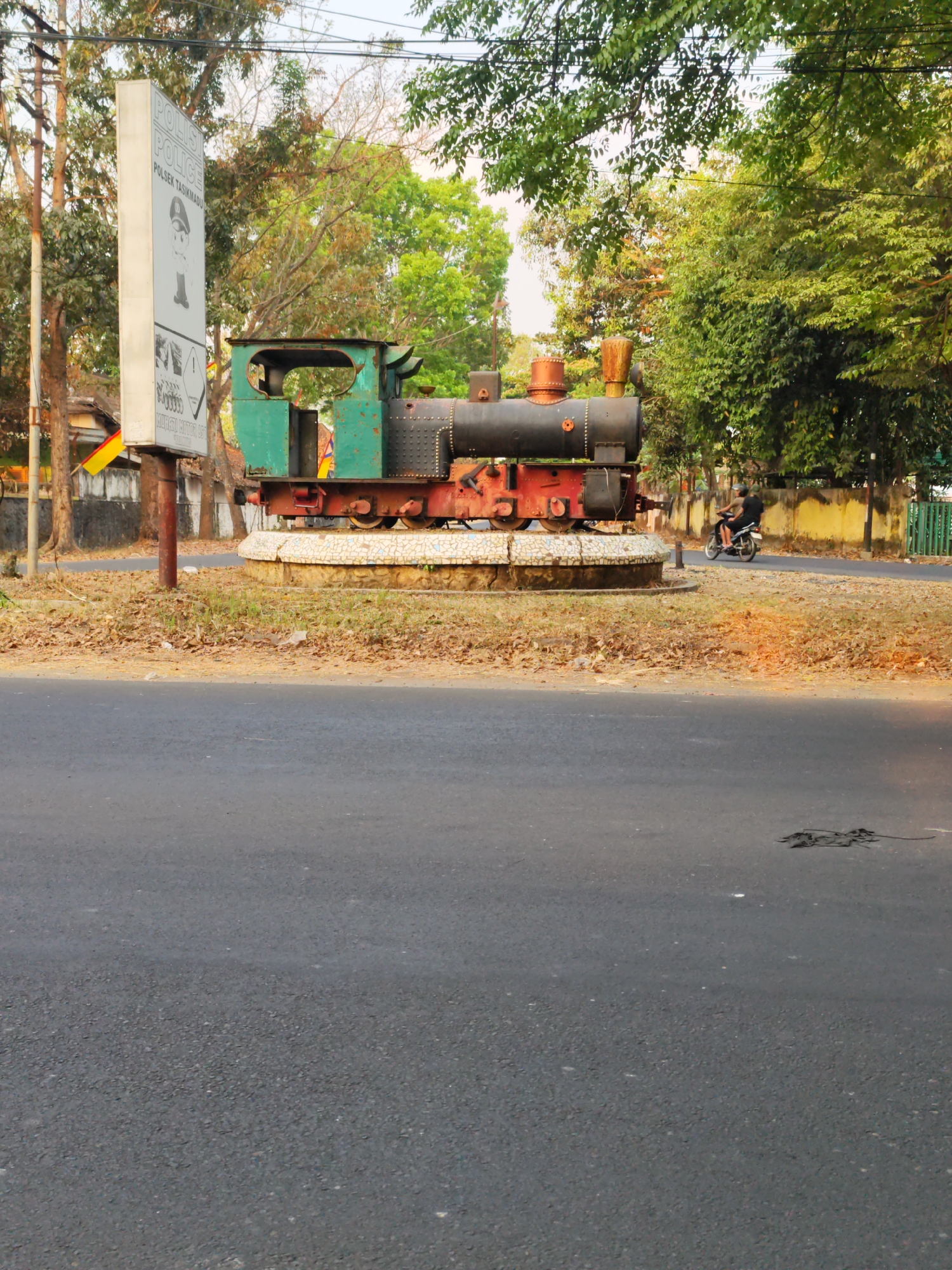 The image shows a stationary, disused locomotive on a circular, tiled base in Tasikmadu, Indonesia. The locomotive is predominantly black and green, with a rusted red chassis and wheel assembly. A tall, white sign displaying the word "POLISI" (police) and the Indonesian text "POLSEK TASIKMADU" is visible to the left of the train. Behind the train, a line of trees with dense green foliage forms a natural backdrop. To the right of the train, a yellow wall and a green metal fence are visible. A person is riding a motorcycle on the road in the middle ground, moving away from the camera. The foreground consists of an asphalt road. The overall scene suggests an outdoor environment in a town or city, likely on the side of a road.
