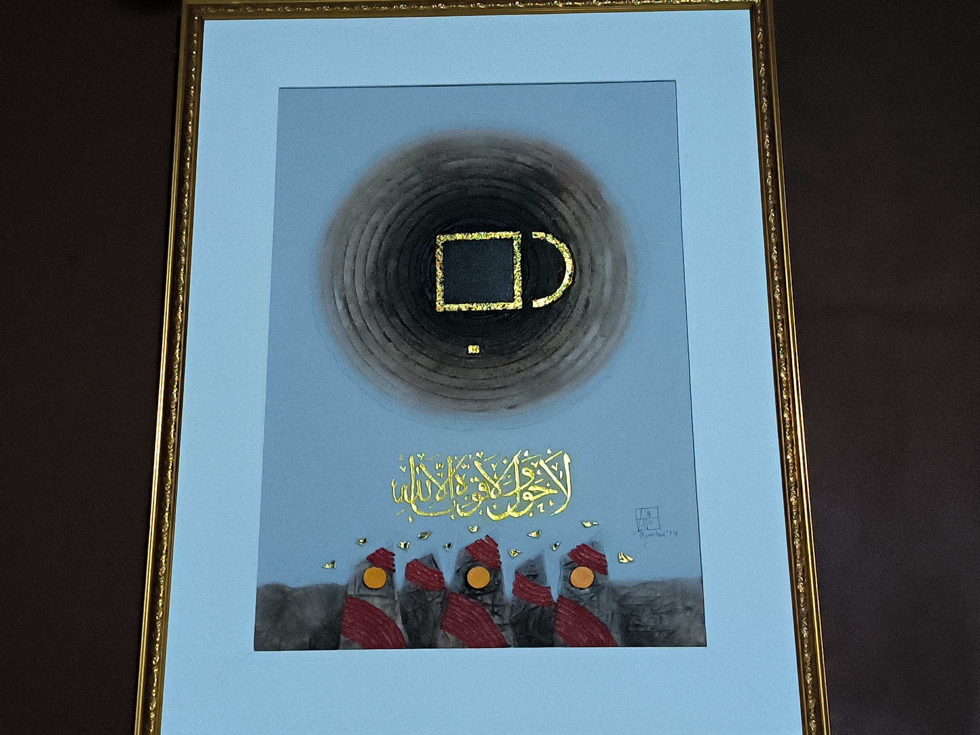 A framed piece of art. The art depicts a black box with gold detailing, enclosed in concentric rings, also of gold. Below, there is gold Arabic calligraphy and five figures with red head coverings and orange faces. Gold leaf flakes are scattered around the calligraphy and figures. The background of the artwork is a light blue color. The frame is gold with ornate beading. The surface the artwork is displayed on is dark brown. The artwork is by Mumtaz '74. This piece of art depicts the Kaaba in Mecca, Saudi Arabia. The city of Lhokseumawe, Indonesia, is not depicted in the image. There are no people in this scene.