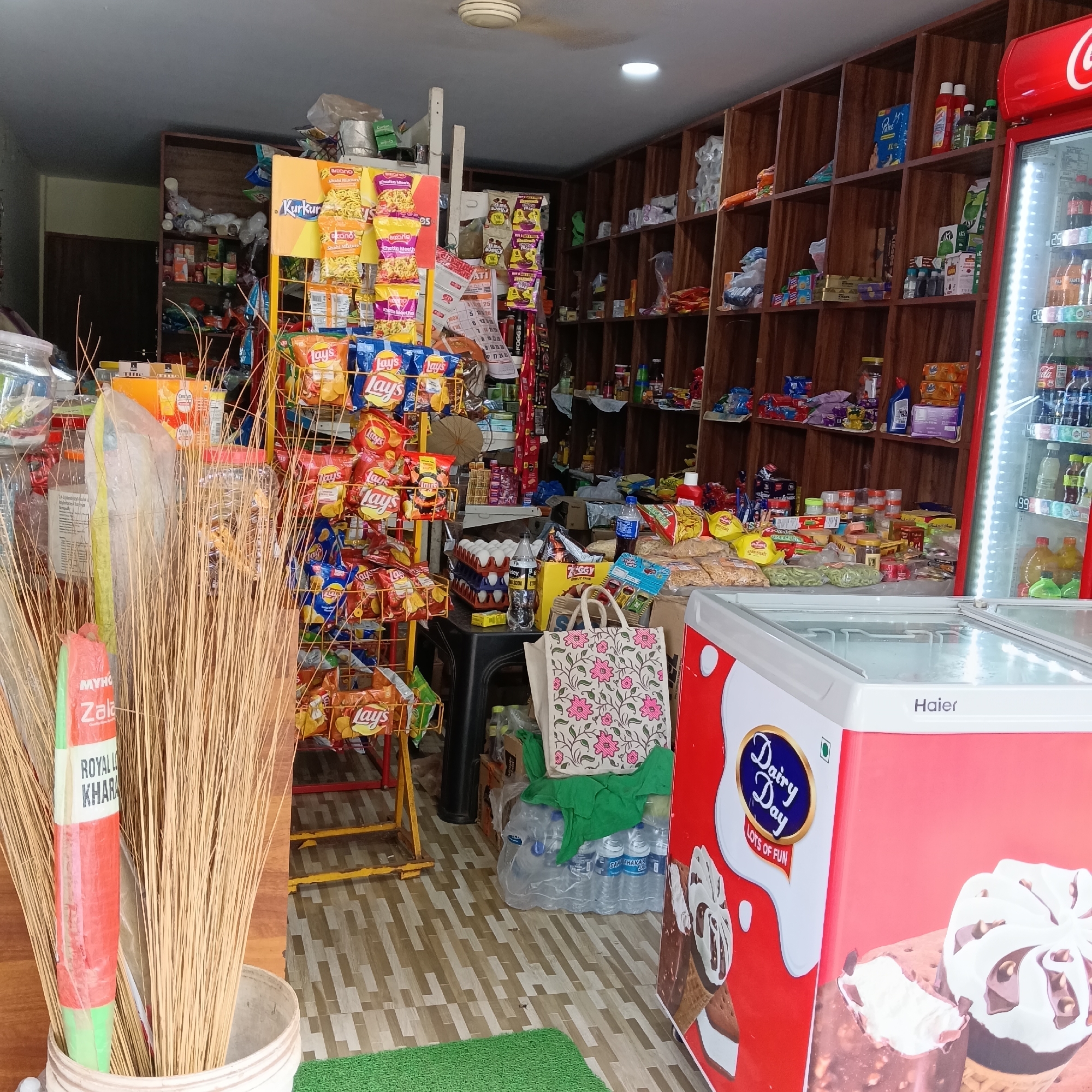 The image captures the interior of a small retail store in what can be inferred as Oxel, India. Dominating the foreground is a bunch of brooms in a bucket on the left, partially obscuring a metal display rack packed with various flavors of Lay's potato chips and Kurkure snacks. Behind the snacks, there is a black table filled with eggs, Ziggy snack box, and a shopping bag adorned with floral patterns.
To the right, a red and white freezer, bearing the brand name "Haier" and "Dairy Day" logo, is visible. The interior shelves of the store are densely stocked with a wide array of products, including snacks, household goods, and groceries. A Coca-Cola refrigerator can be seen on the right side.
The overall impression is that of a compact, well-stocked local store with a focus on snack foods and chilled items. The store exhibits the character of a typical small business found in urban India.