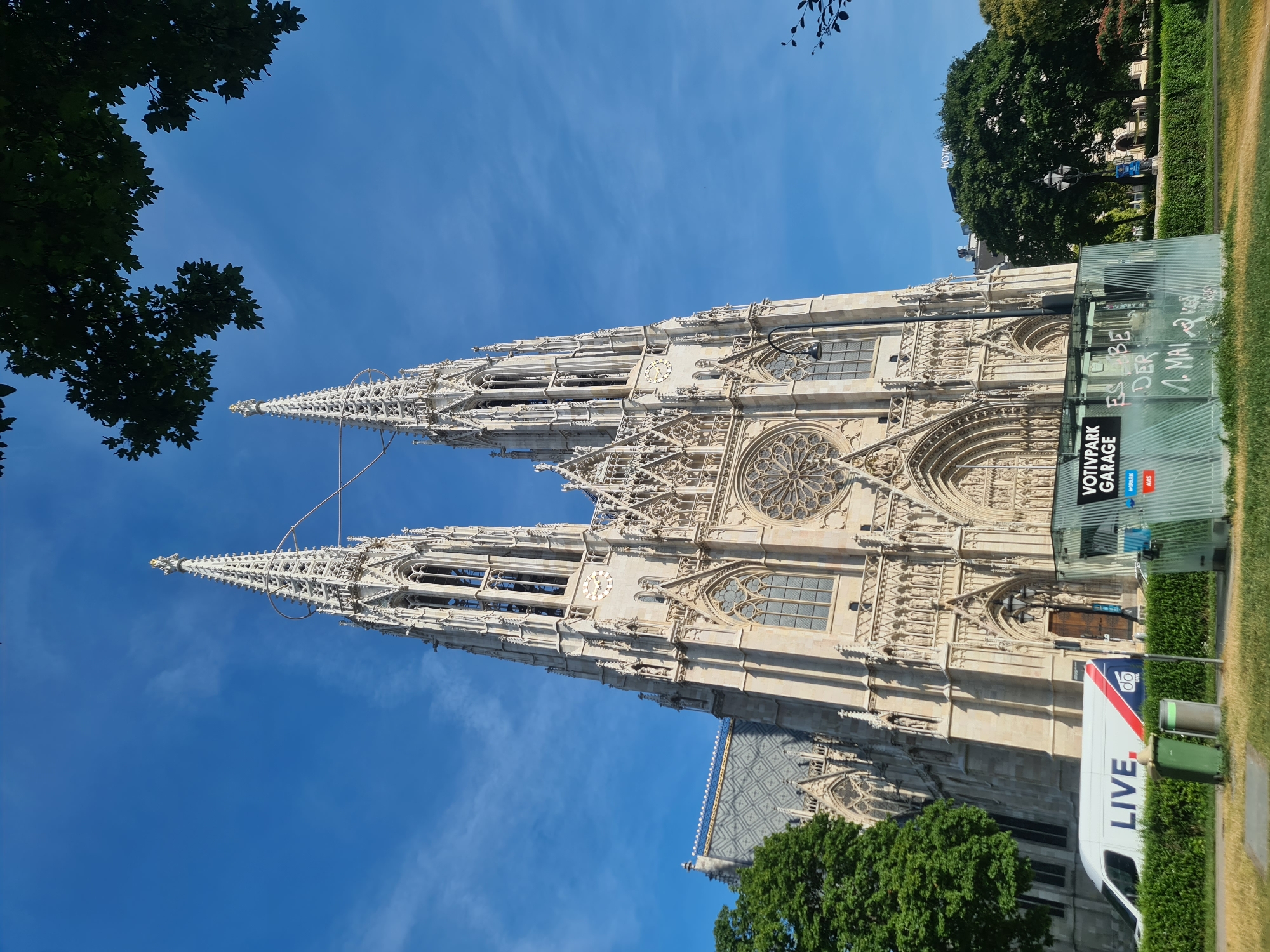 The Votivkirche in Vienna, Austria, is depicted under a clear blue sky. The Gothic Revival church features two tall spires, intricate stone carvings, and a large rose window. In the foreground, a white van with "LIVE" written on its side is parked in front of a green hedge. To the right of the van, a temporary structure with the words "VOTIVPARK GARAGE" is visible, adorned with graffiti. Trees frame the scene, with lush green foliage on the left and darker leaves from a higher branch casting shadows from the top right. The overall scene captures the grandeur of the church juxtaposed with modern elements and urban greenery.