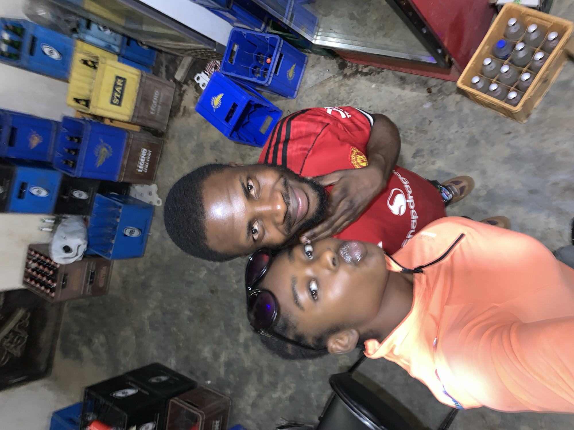 The image appears to be a selfie taken indoors. Two people are present: a man wearing a red soccer jersey with black stripes and a woman in an orange top with sunglasses perched on her head. They are posing close together and smiling. The background includes numerous stacked plastic crates, some labeled "STAR" and "LEGEND," along with a crate of glass bottles. Based on the mention of "New Stadium, Nigeria," the image was likely taken at or near a location selling beverages or other goods within the area.