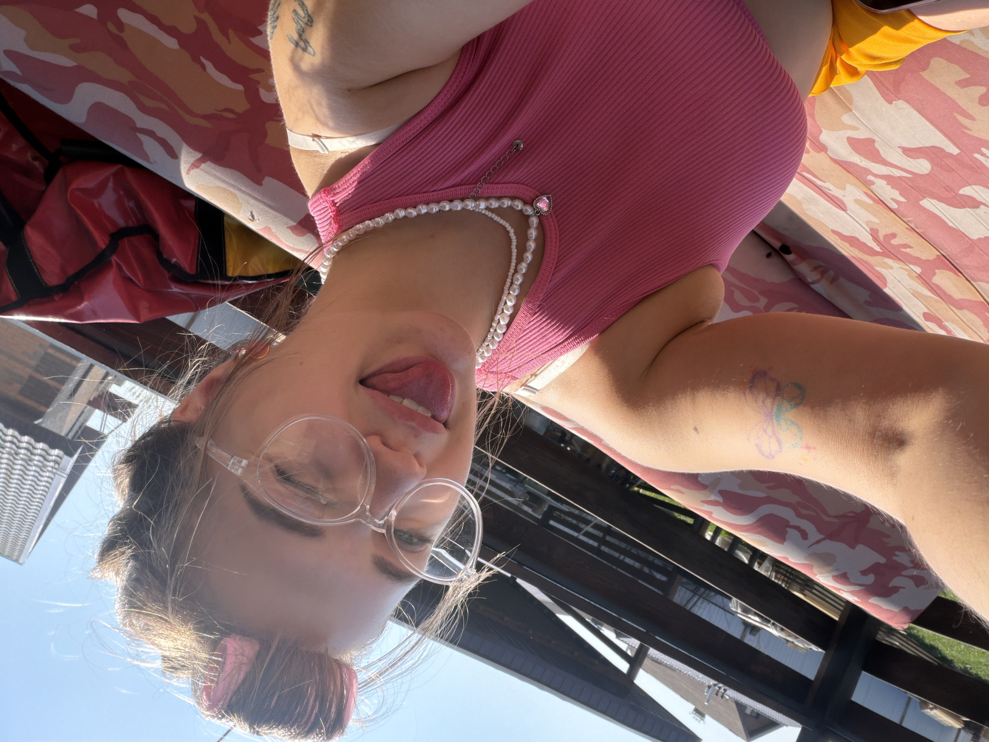 A woman wearing pink glasses and a pink top with a pearl necklace is sticking her tongue out. She has a hair clip in her hair and a tattoo on her arm. She is sitting on a pink and white camouflage print surface, possibly at an outdoor location in Большая Шильна, Russia, based on the location metadata. A building and a structure with horizontal beams are visible in the background.