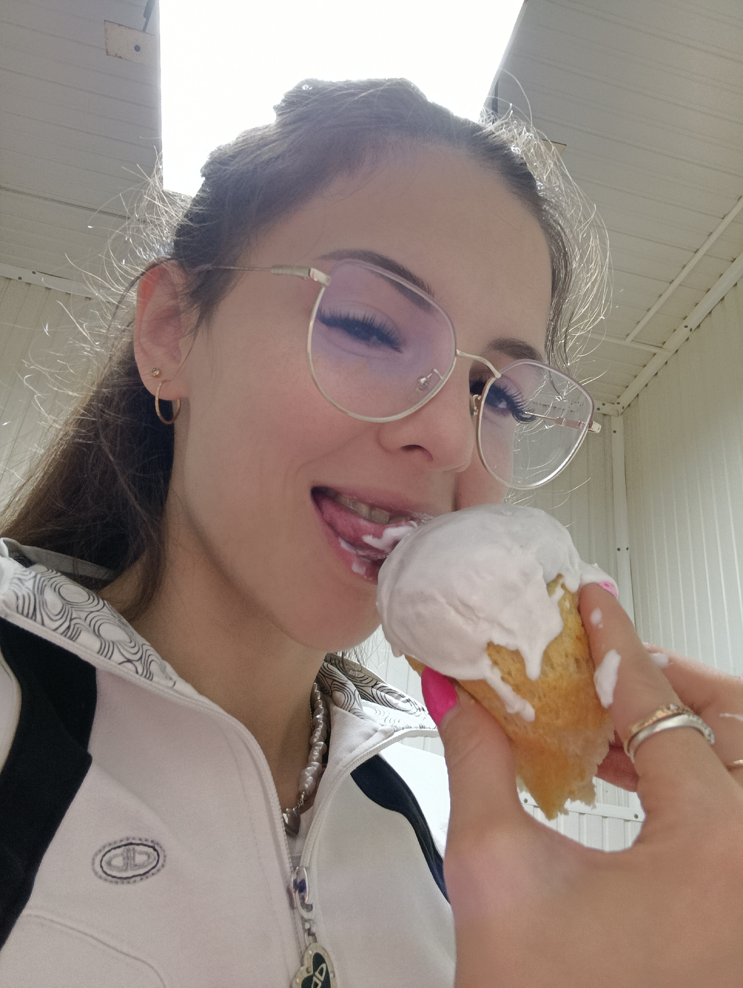 A woman with long brown hair and gold-rimmed glasses is eating a cake with white frosting. She is wearing a white jacket with black accents and a necklace. The woman is wearing two rings on her hand, and her nails are painted bright pink. The background is a white wall with vertical lines. The location is Прикамский, Russia.