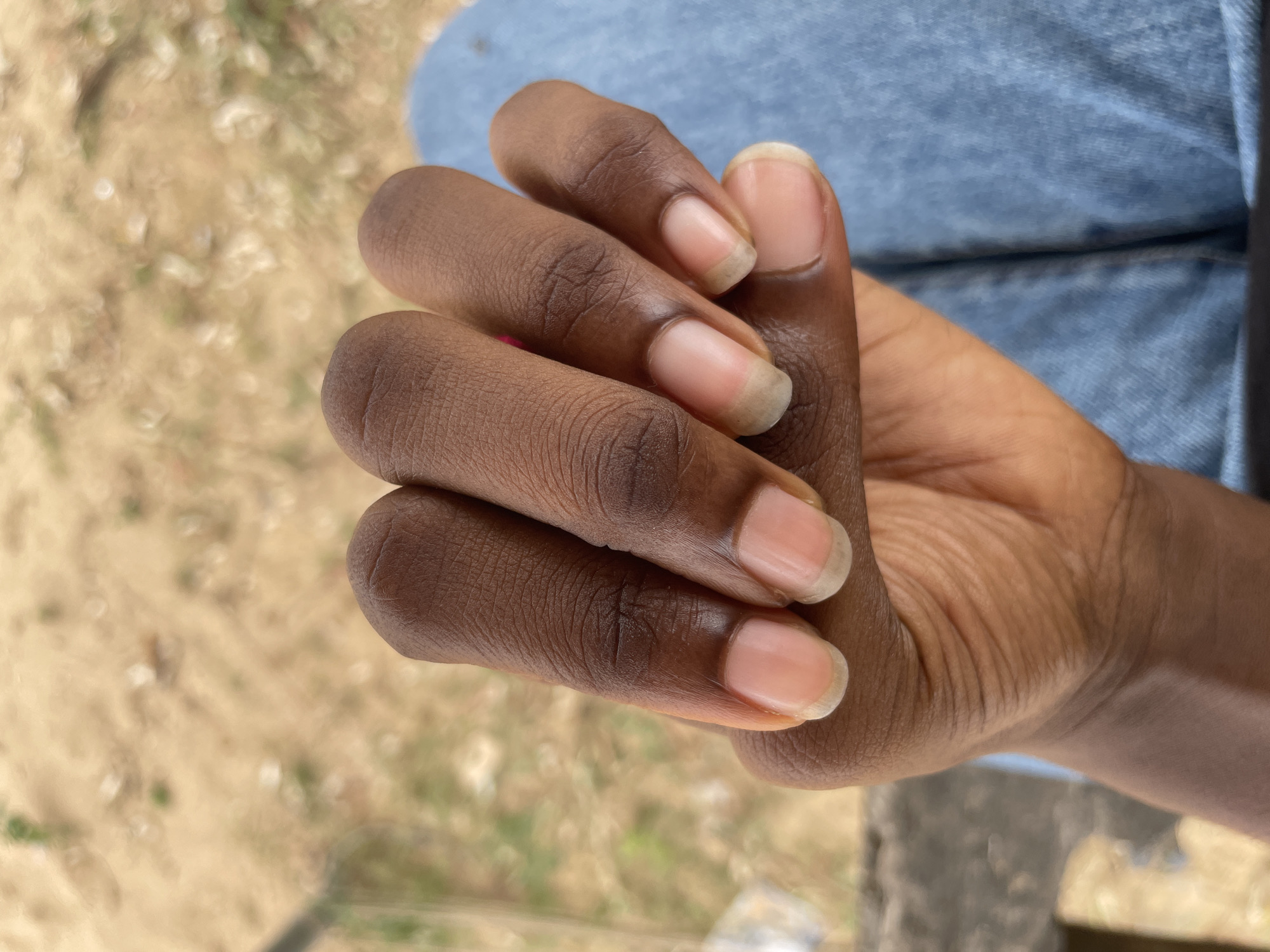 A close-up, overhead shot shows a person's right hand, with fingers curled inwards, resting on a pair of blue denim jeans. The hand has dark skin with visible texture and lines on the knuckles and fingers. The fingernails are short and naturally shaped, appearing clean and healthy with a slightly pinkish hue. The background is blurred, showing a sandy, earthy surface with patches of dried grass and some out-of-focus greenery, suggesting an outdoor setting. The overall impression is of a casual, everyday moment. The scene is located in Iyafin, Nigeria.