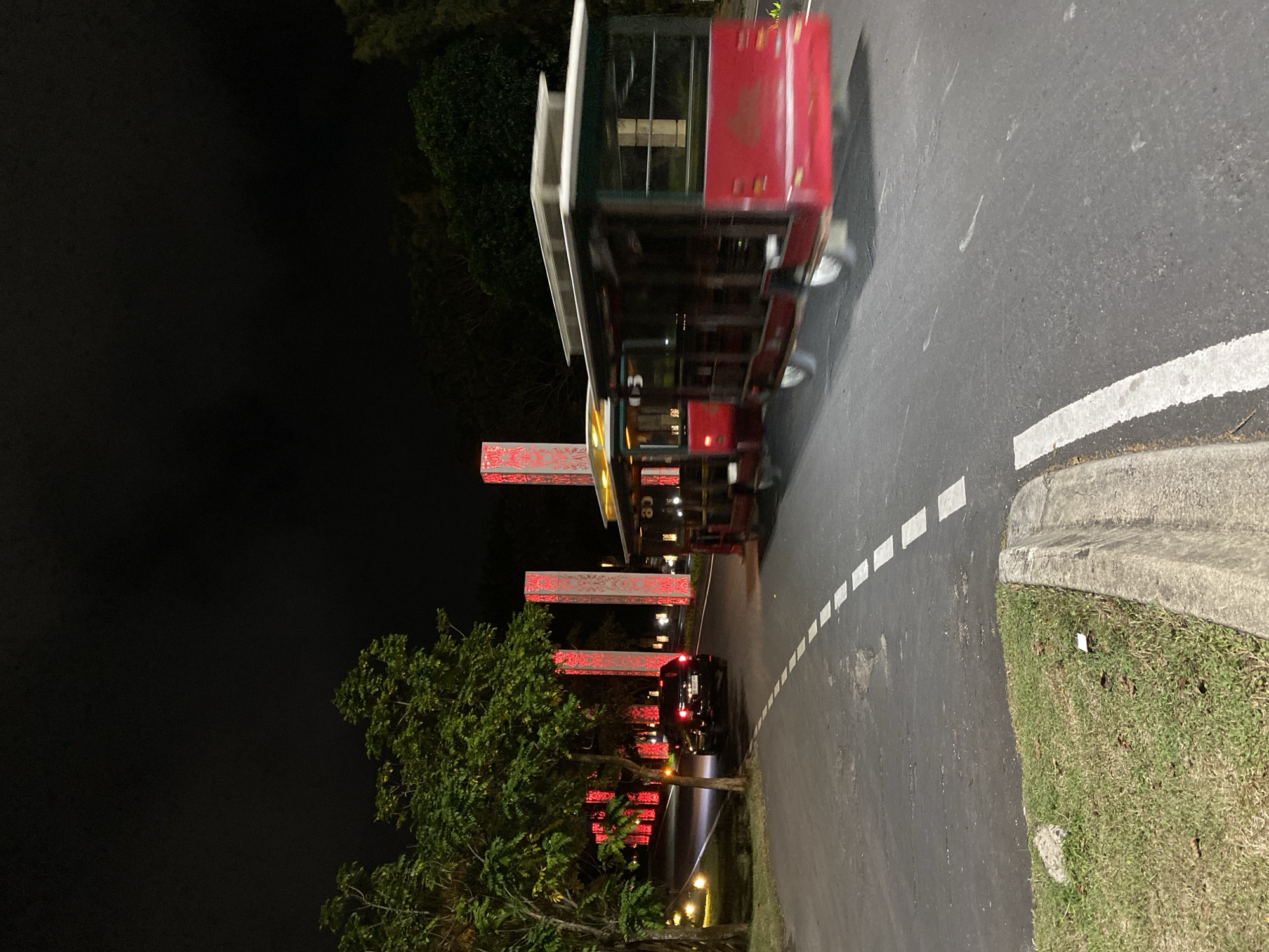 In the city of Kuta Selatan, Indonesia, at night, a red and white open-air trolley, possibly a tourist shuttle, is captured in motion on a paved road. The trolley's wheels are blurred, indicating movement. Adjacent to the road, a black SUV is parked on the left side. The environment is illuminated by strategically placed red lights emanating from tall, decorative pillars that line the road, creating a vibrant and festive atmosphere. Lush green trees are visible on the left, with their leaves illuminated by ambient light. The road itself is marked with dashed white lines. The sky above is dark and clear.