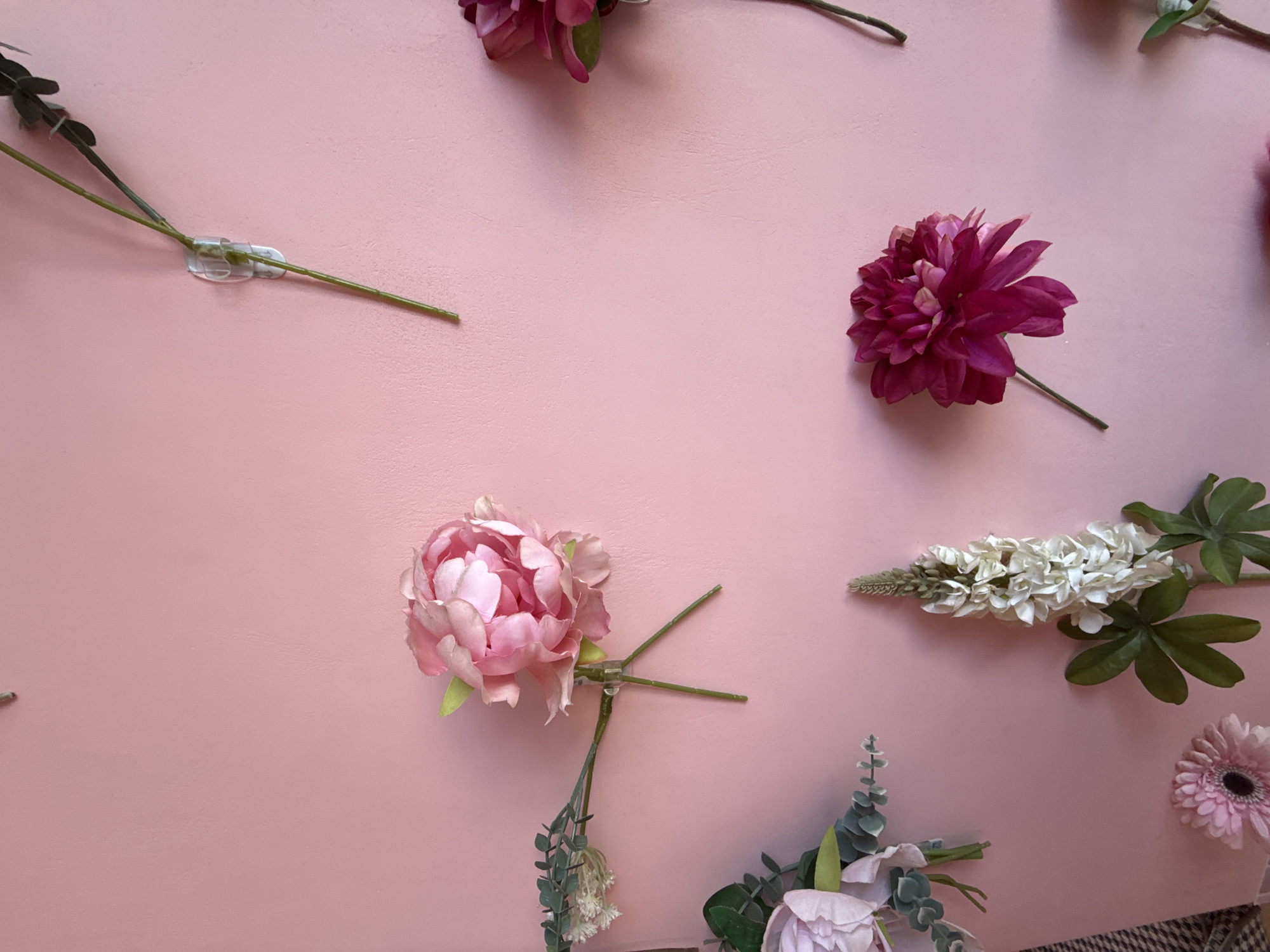 The image shows a close-up of artificial flowers attached to a pale pink wall using clear adhesive hooks. The flowers are of various types and colors, including pink peonies, deep pink dahlias, white lupines, and light pink gerbera daisies. Some of the flowers are in full bloom, while others are still buds or have smaller accompanying foliage. The lighting is even, suggesting an indoor setting. There is no indication of location or people in the image.