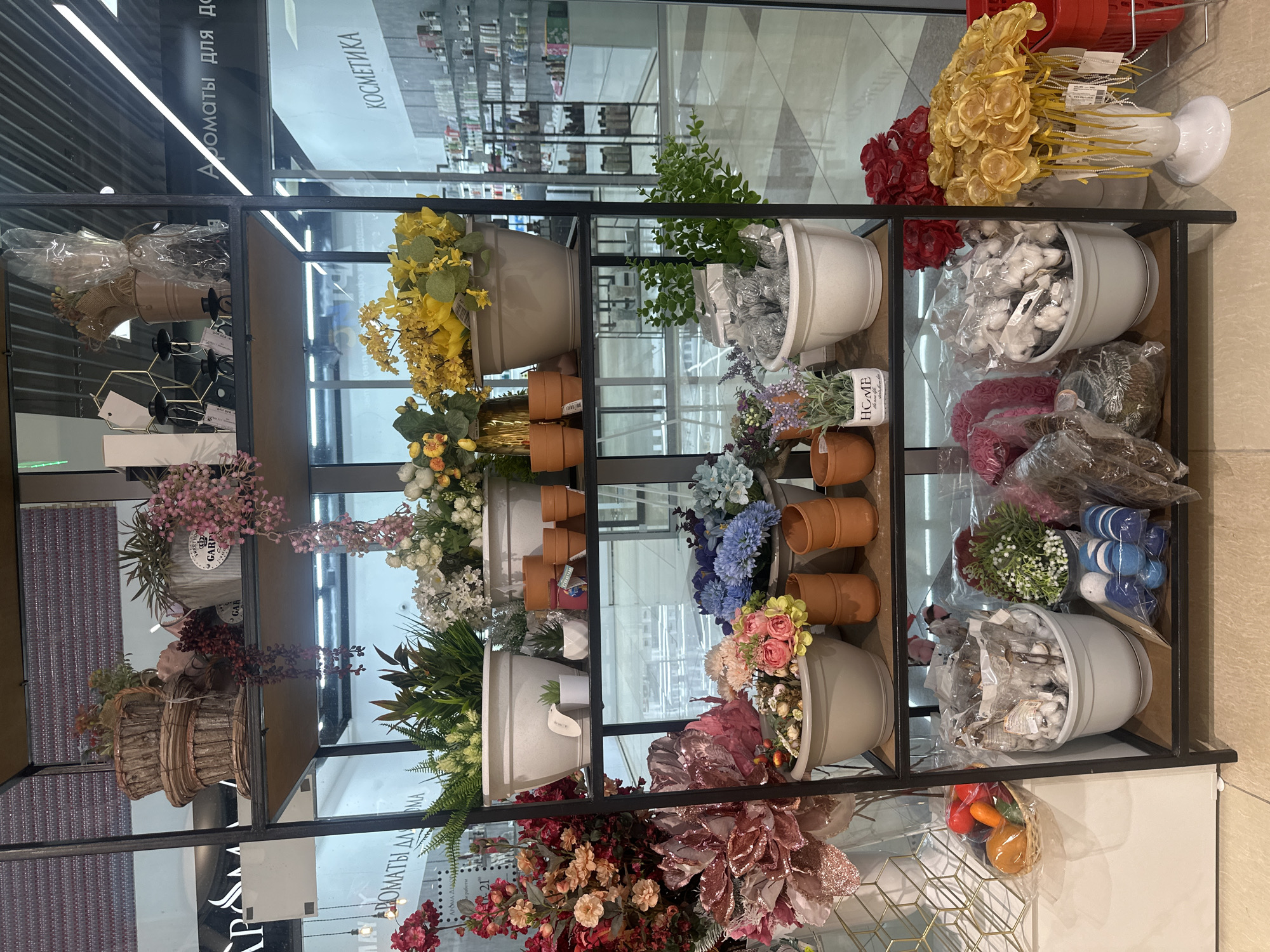 The image shows a retail display shelf filled with artificial plants, flowers, and decorative items in a store. The shelf is constructed of black metal with wooden shelves. On the top shelf, there are stacked woven baskets with plants, a decorative pot with pink flowers, and a gray pot with a "Garden" label. The second shelf features large white planters with various green plants, a row of terracotta pots, and a gold vase with yellow flowers. The third shelf displays a large pot with pink and red artificial flowers, a smaller pot with pink and blue flowers, and several terracotta pots. The bottom shelf contains a white pot filled with cotton balls in plastic packaging, a pot with green foliage, a basket of small artificial fruits, and a white vase with yellow decorative flowers. In the background, shelves of products are visible, suggesting a store setting. The overall impression is one of a well-stocked floral or home decor section of a shop. The text visible in the background appears to be in Russian, with phrases like "АРОМАТЫ ДЛЯ ДО" and "КОСМЕТИКА," which translate to "AROMAS FOR THE HO" and "COSMETICS." This indicates the store is likely a cosmetics or home goods store. Given the Russian text, the location can be inferred to be in Russia, and specifically, it is identified as Petropavlovsk-Kamchatsky, Russia, based on the provided context. There are no people visible in the image.