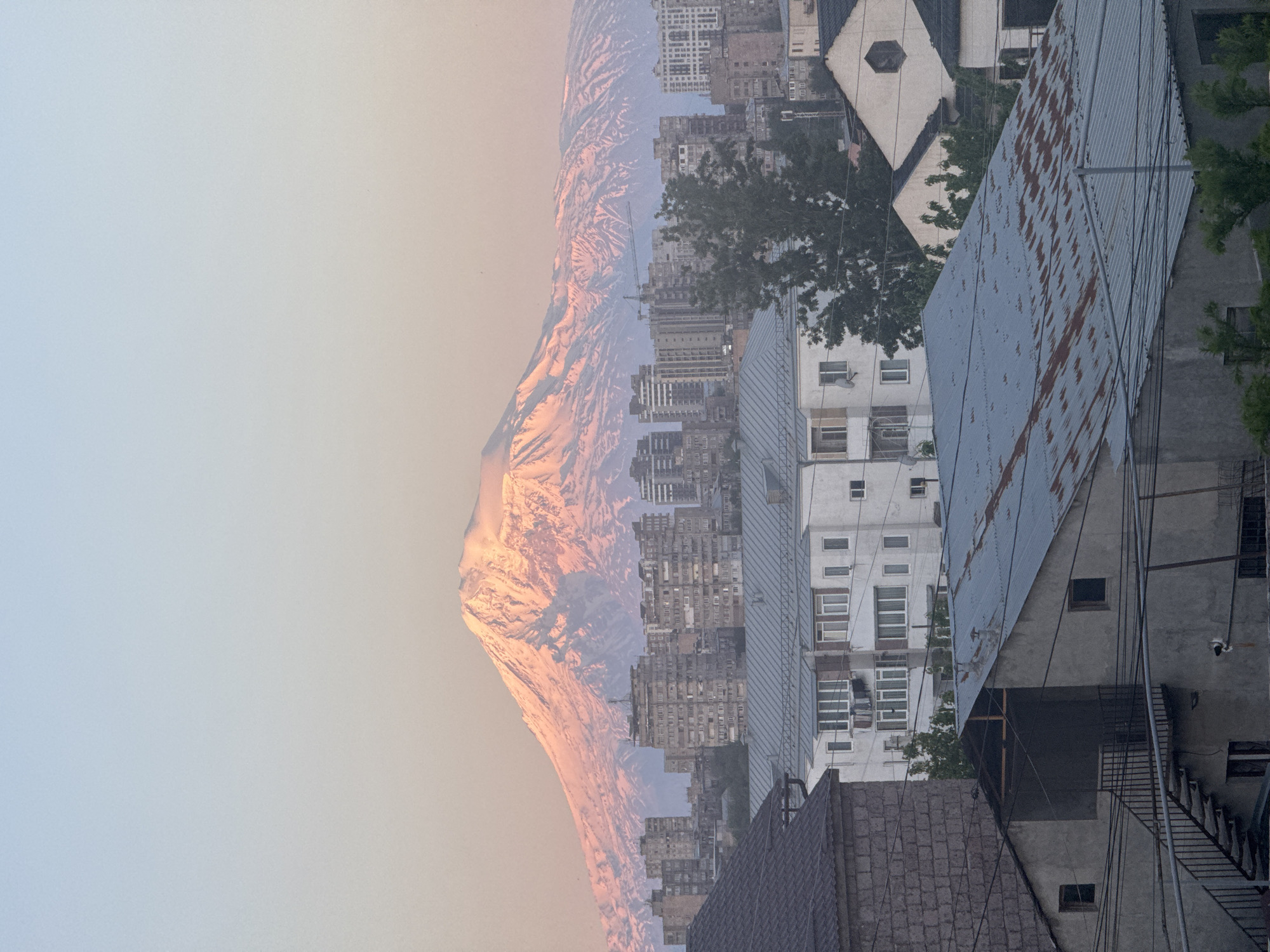 An image showing a cityscape with a mountain in the distance, likely taken in Yerevan, Armenia. The mountain, presumably Mount Ararat, is to the left, with its peak covered in snow, bathed in a soft, warm light. In the foreground are buildings of varying heights and architectural styles. Some have flat roofs and multiple windows, while others have angled metal roofs, with signs of rust or corrosion. Power lines are visible, running across the scene, intersecting the buildings. The sky above the mountain is light, with a gradient of light blue to pale orange near the horizon.