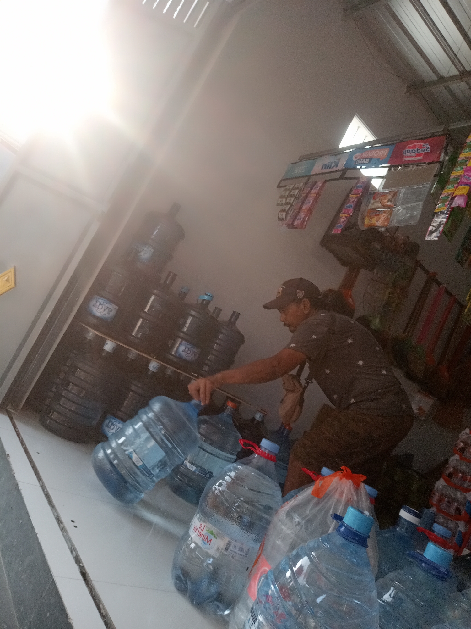 A man wearing a baseball cap, dark t-shirt with a leaf pattern, and patterned shorts stands in a shop in Simo, Indonesia. He is reaching for a large, clear plastic water jug with a blue cap, which is partially obscured by other water jugs in the foreground. The shop appears to be a small convenience store or warung, with shelves stocked with various packaged goods hanging on the wall. The floor is tiled, and bright sunlight streams in from an open doorway or window on the left side of the frame, creating lens flare. A large stack of dark-colored water jugs is visible in the background. The overall impression is a scene of daily commerce in a small Indonesian town.