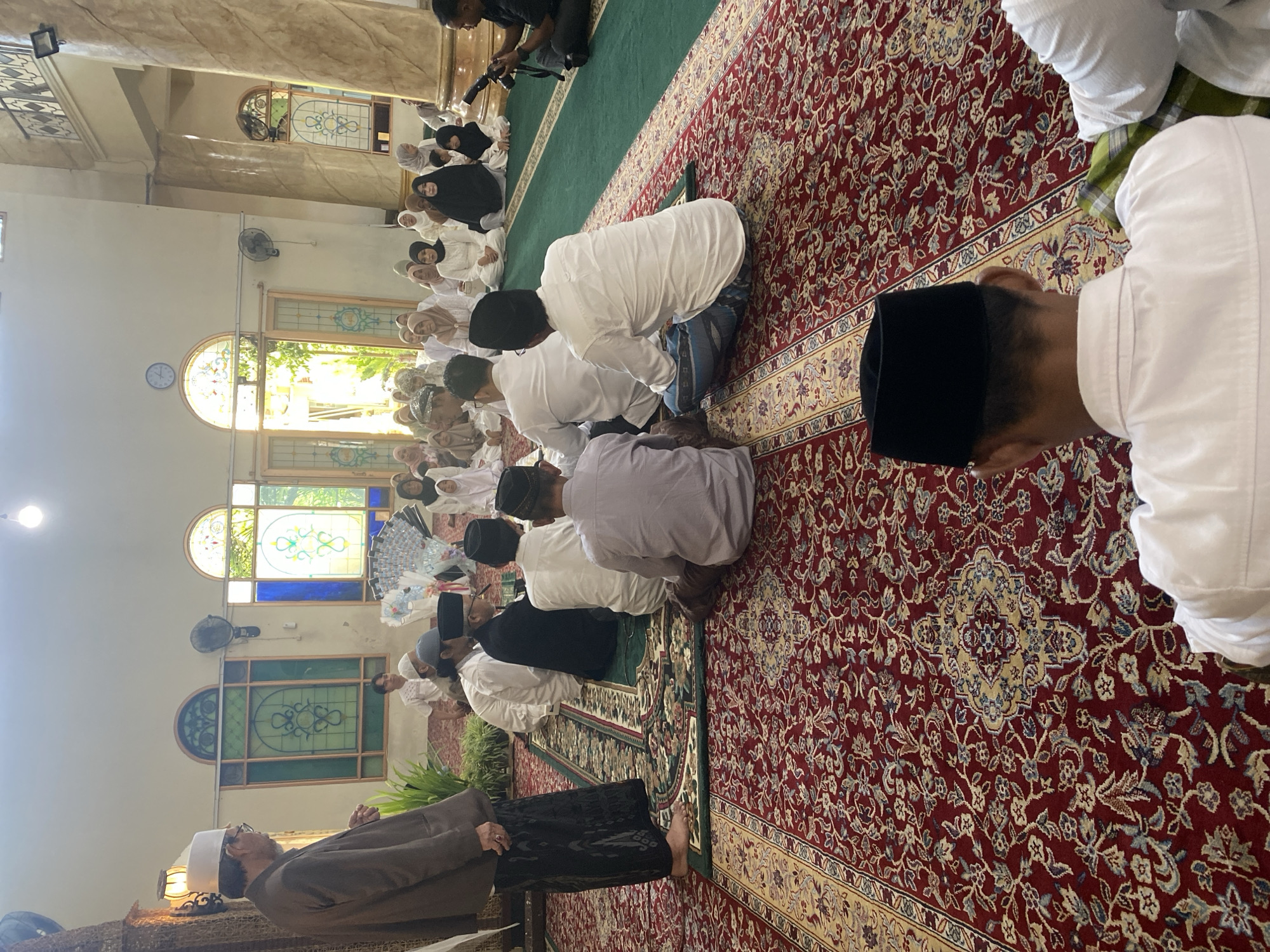 A group of men and women are seated on a red carpet inside a room with an ornate patterned floor. The room features stained-glass windows and tall marble columns. One man, dressed in a brown suit and a white peci (hat), stands on the left side of the image, facing the seated individuals. Several men in white shirts and peci hats are seated in the foreground, facing away from the camera. In the background, men and women are also seated, many wearing headscarves. A person on the right is holding a camera, appearing to film the gathering. The scene suggests a religious or ceremonial event taking place in Bondowoso, Indonesia, likely within a mosque or a similar communal space.