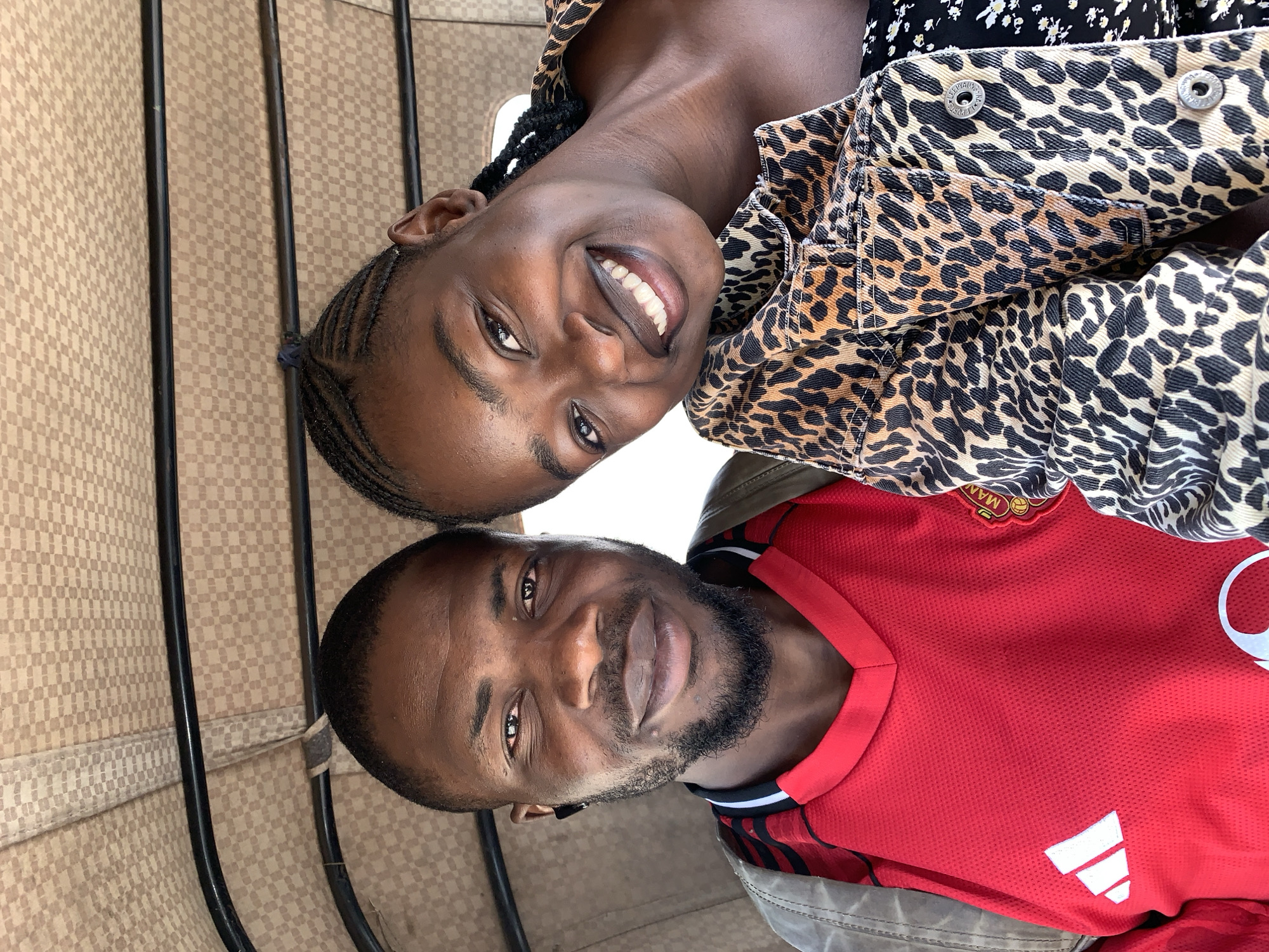The image, when viewed upright, captures a man and a woman posing closely for a cheerful photo, likely a selfie. Both individuals have dark complexions and are smiling.
The woman, positioned at the top of the frame, looks directly at the camera with a broad, joyful smile, revealing her teeth. Her hair is styled in dark braids. She is wearing a striking leopard-print jacket, possibly denim, with metallic buttons visible near the collar.
Below her is the man, also looking towards the camera with a pleasant, subtle smile. He has a short beard. He is dressed in a bright red sports jersey with a V-neck, featuring a white Adidas logo on the chest. Above the Adidas logo, a football club crest is partially visible, showing the letters "MAN" and hinting at Manchester United.
The background suggests they are inside a vehicle, possibly public transport like a tricycle (Keke Napep) or a bus. The interior is upholstered with a light brown, repeating checkerboard pattern. Several vertical black bars or poles are visible behind them. The bright lighting indicates it is daytime. Both subjects convey a happy and relaxed mood. The location context for this scene is Rafin Par, Nigeria.