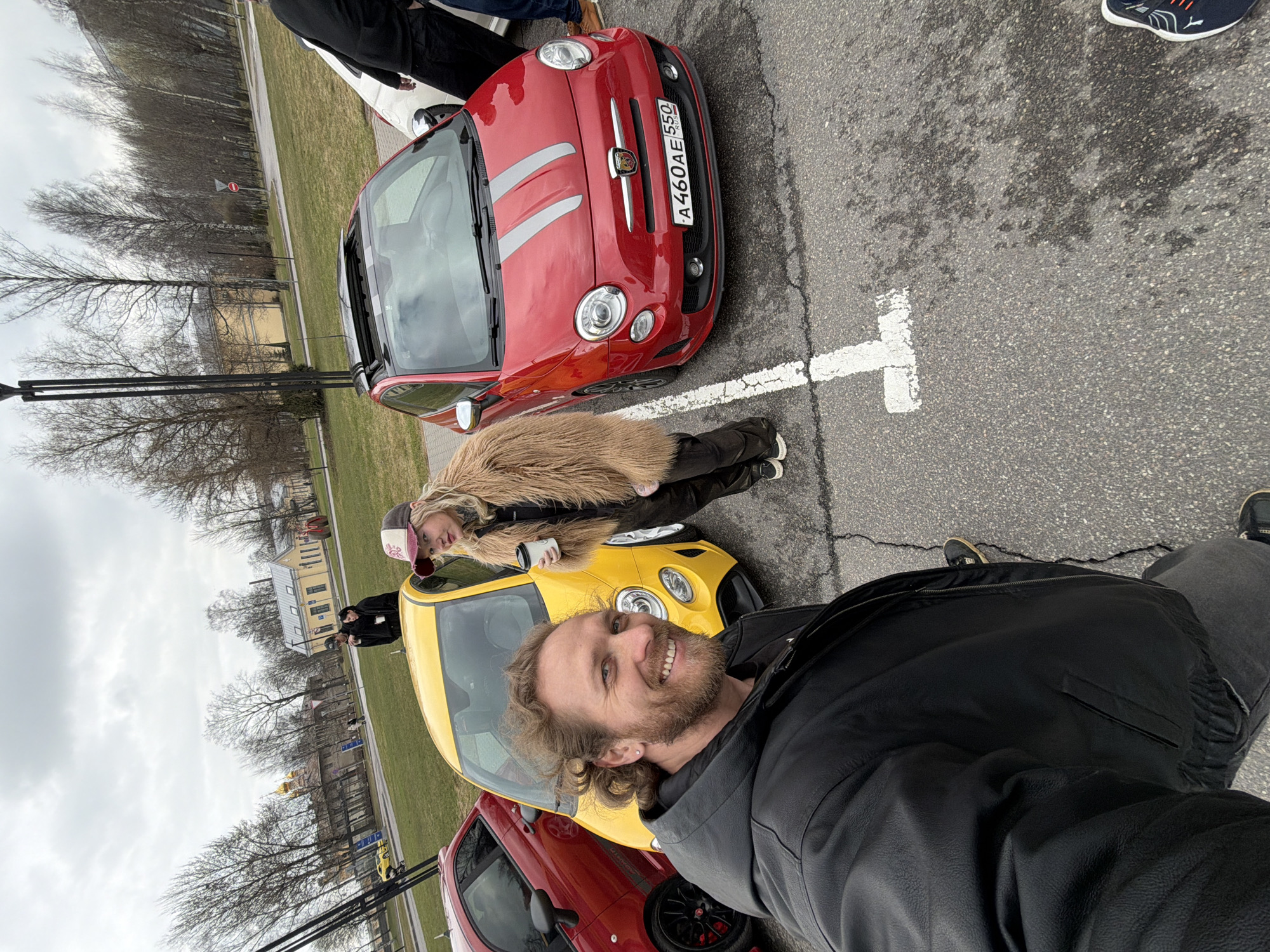 The image captures a man taking a selfie with two women in a parking lot in Saint Petersburg, Russia. There are four cars visible: two red Fiat 500 Abarth models, and one yellow Fiat 500. The red car directly behind the photographer has a white stripe down the middle. The license plate of the front red car reads "A460AE 550 RUS". The second car in the background is yellow. The woman on the left is wearing a large fur coat and holding a white paper cup. She is wearing a baseball hat. The lot is marked with painted white lines to indicate parking spaces. In the background there is grass and various buildings, including an old two story building, and a building with yellow walls. There are several leafless trees. The sky appears overcast.