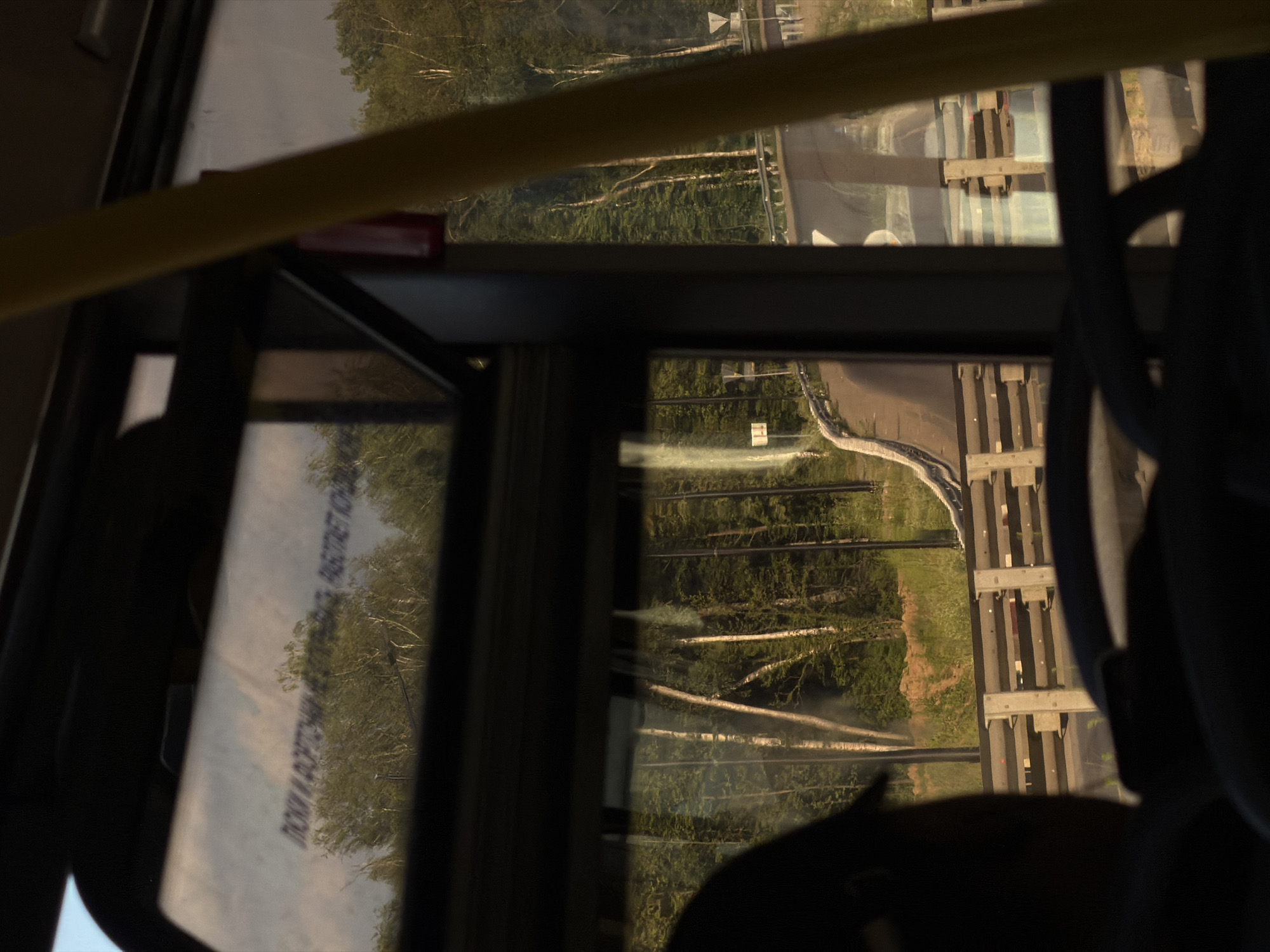 The image depicts a view from inside a vehicle, likely a bus or train, looking out of a window. The scene outside is dominated by a lush green forest with numerous trees, some of which appear to be birch trees with their characteristic white bark and branches. The ground is covered in grass and vegetation.
A winding road or path is visible in the middle distance, with a low concrete barrier along its edge. Further in the background, there are structures that resemble railway tracks or an elevated walkway with wooden and metal supports. The reflection on the window suggests that the photo was taken during daylight, likely late afternoon or early evening, given the warm, golden light illuminating the scene.
The interior of the vehicle is partially visible, with dark seats and a yellow handrail in the foreground, indicating a passenger's perspective. The window has some text printed on it, which is partially visible and appears to be in Cyrillic script. The text is distorted by the reflection and the angle, making it difficult to read clearly, but it might be instructional or informational.
The overall impression is one of a serene journey through a natural landscape, with the contrast between the man-made interior and the natural exterior creating a sense of peaceful observation. The lighting and reflections suggest a calm and pleasant atmosphere.