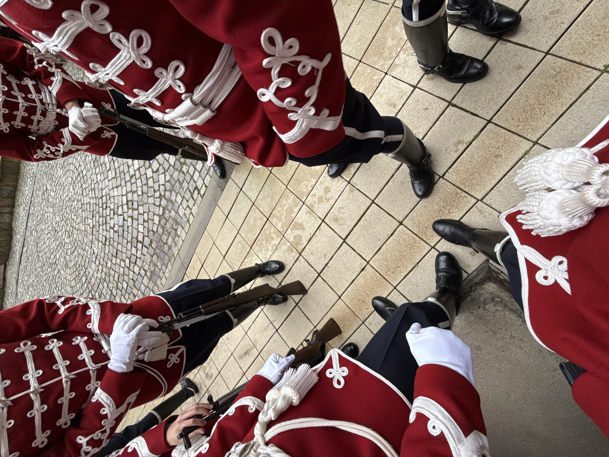 The image shows a group of Bulgarian soldiers standing in formation in Sofia, Bulgaria. The soldiers are wearing red uniforms with white decorative trim, black trousers with a white stripe down the side, and black boots. They are holding rifles with bayonets attached. The ground is paved with tiles and cobblestones.