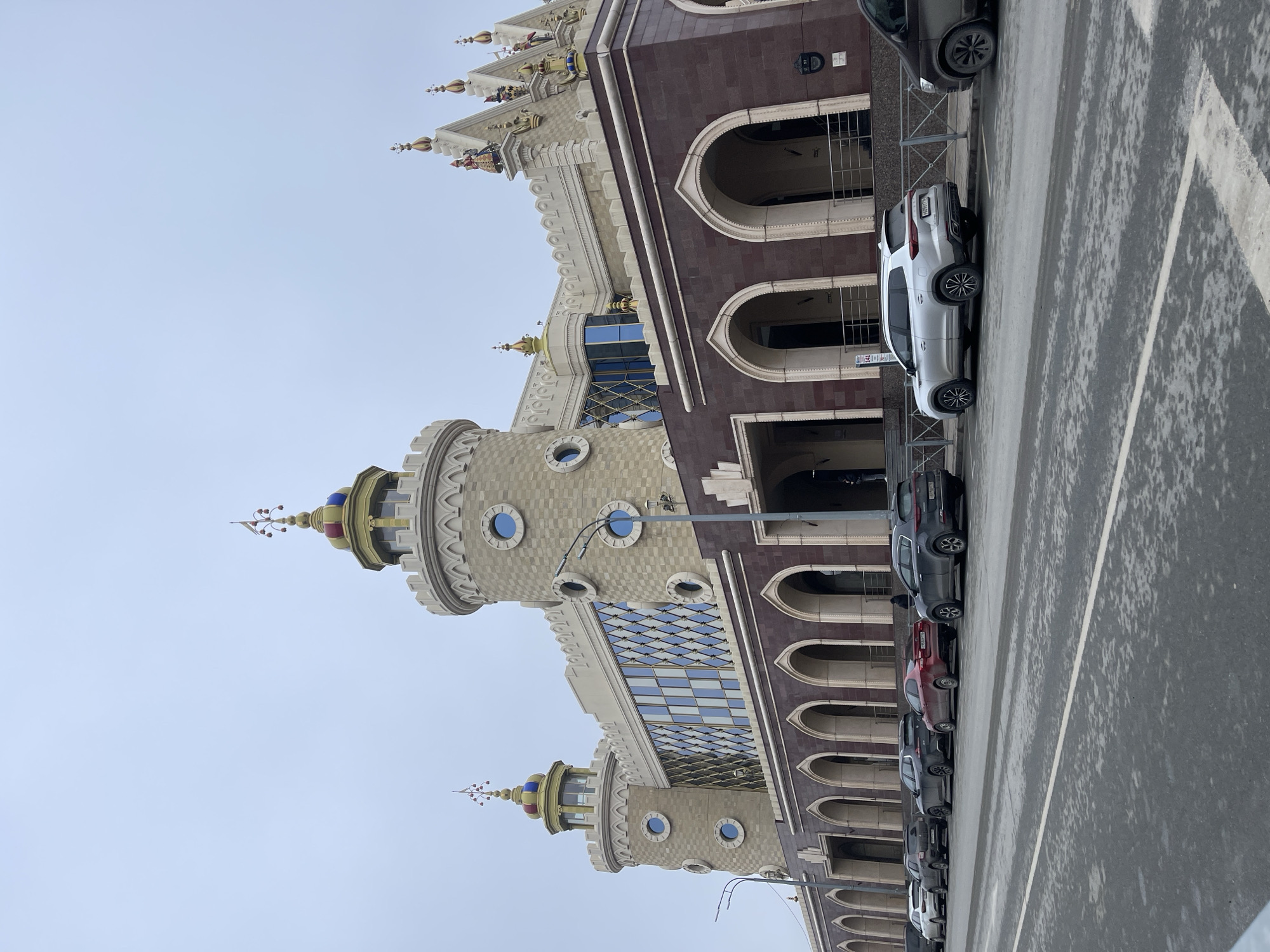 The image captures a view of the Kazan Puppet Theatre "Ekiyat" in Kazan, Russia. The building's architecture features towers with round windows and decorative elements. A row of parked cars lines the street along the building. The sky is overcast. There are no visible people in the frame. The setting includes a paved street with parking lines.