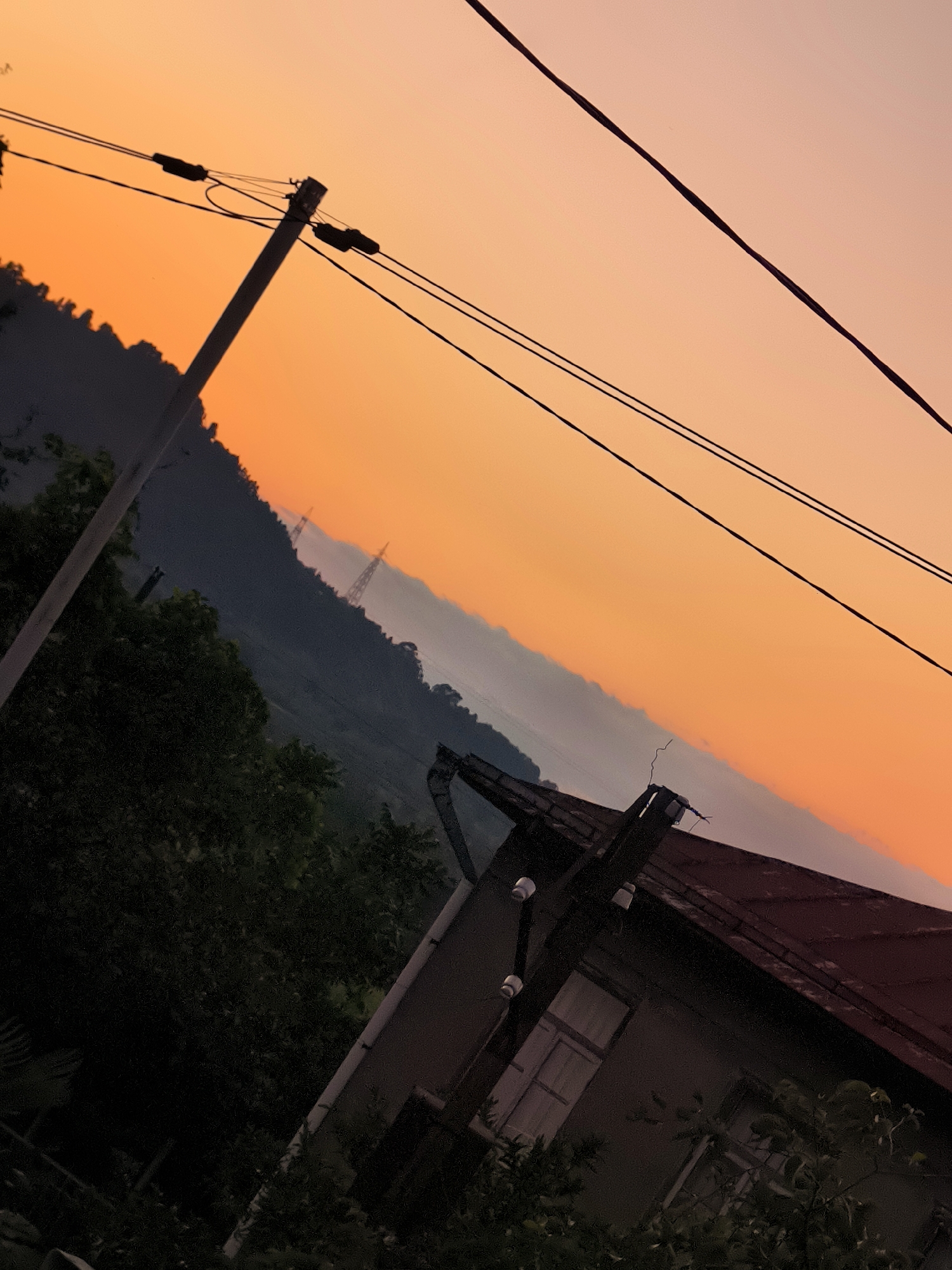 The image captures a landscape scene at sunset in Makho, Georgia. The sky is a gradient of orange and pink, suggesting the setting sun. In the foreground, a dark silhouette of trees and a portion of a building with a red roof are visible. A utility pole with wires runs diagonally across the frame. In the middle ground, a hillside is covered with trees, and in the distance, power line towers are visible on a ridge. The overall impression is of a rural or semi-rural setting during twilight.