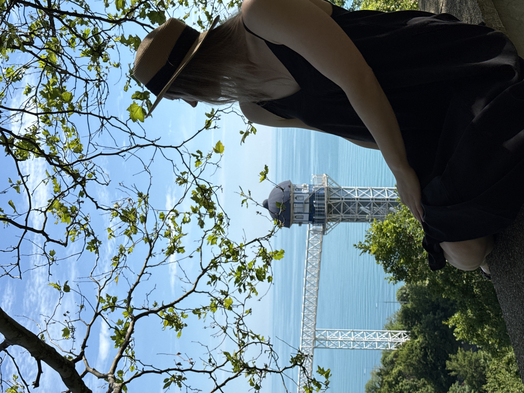 The image depicts a woman in a black dress and a straw hat sitting on a stone ledge, looking out at a scenic view. In the background, a vast expanse of turquoise water stretches to the horizon, under a bright blue sky dotted with wispy clouds. Overlooking the water is a distinctive architectural structure, a tower with a dark domed roof, connected by a white metal bridge-like walkway to another white lattice tower. The scene is framed by the branches of a tree with green leaves, some of which are in the foreground, casting dappled shadows. Lush green trees cover the hillside below the ledge. The location is identified as Nebug, Russia.