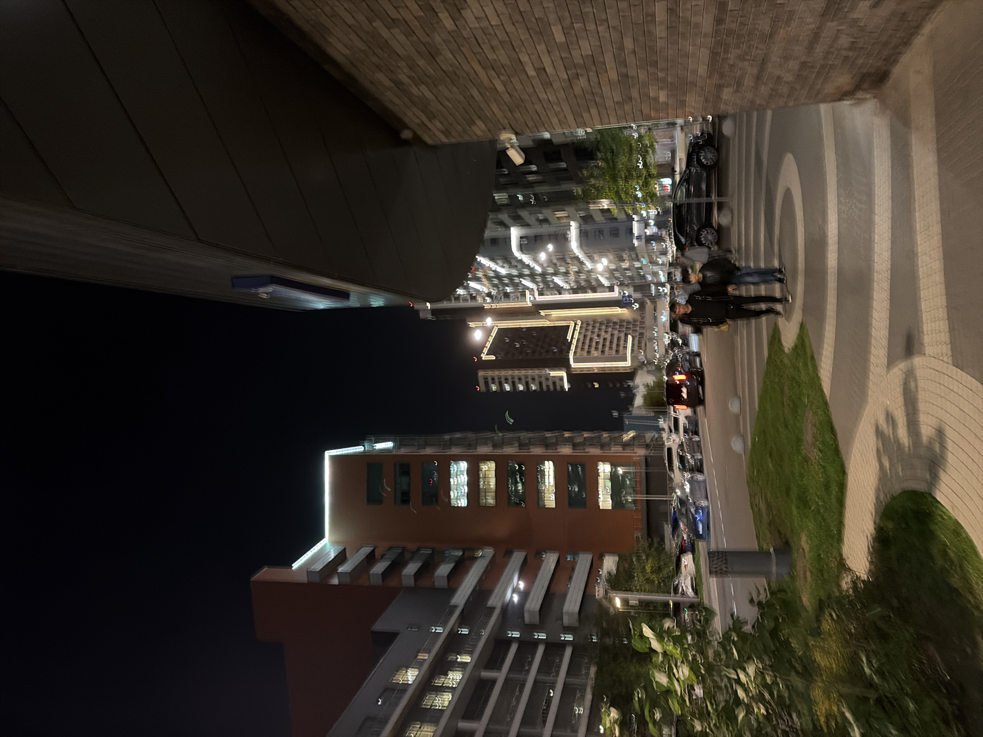 The image depicts a night scene in Moscow, Russia, specifically on a pedestrian walkway and street in a modern urban area. The perspective is from ground level, looking towards a street lined with multi-story buildings.
In the foreground, a paved walkway with a distinctive circular pattern and a bordering grass area is visible. To the right, a textured brick wall forms the side of a building, with a dark, overhanging structure above it.
Two individuals, appearing to be a man and a boy, are walking across the pedestrian area away from the viewer. The man is wearing a dark jacket and pants, and the boy is in a dark jacket and jeans.
The street in the background is illuminated by streetlights and the lights from the buildings. Several cars are parked or driving on the street, including a dark SUV and a dark van. The buildings are modern, with many windows reflecting the night lights. One prominent building on the left is orange-red with several floors of windows and balconies, accented with horizontal white lines. Further down the street, other tall buildings are visible, some with vertical lighting accents. The sky above is dark, characteristic of nighttime. The overall impression is of a well-lit, contemporary urban environment at night.