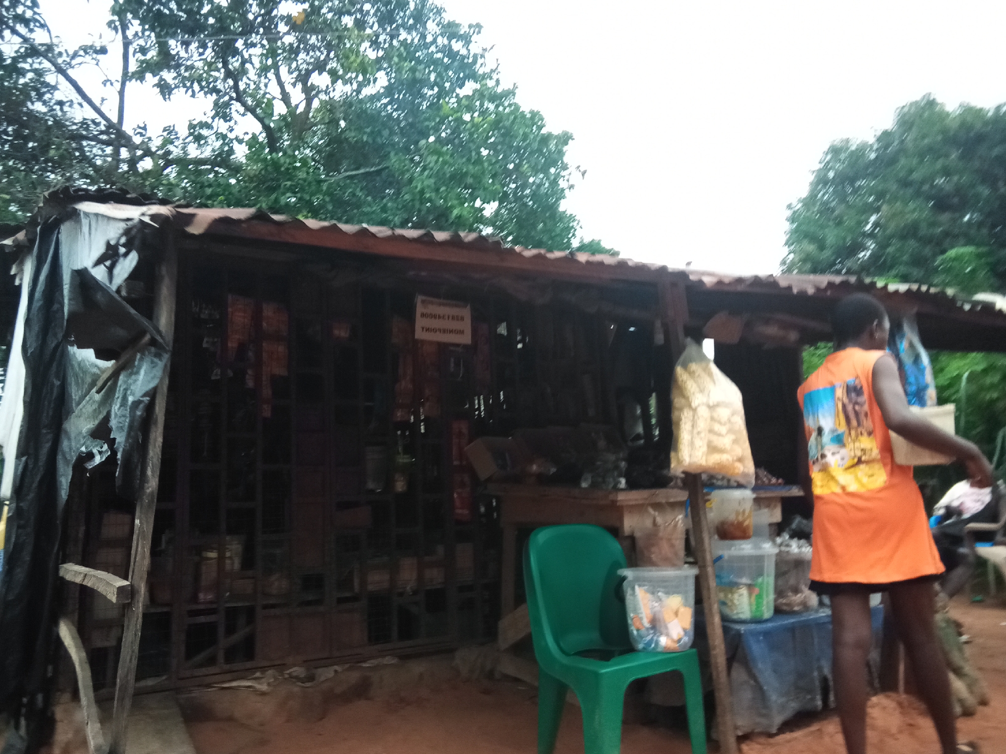 A medium shot depicts an outdoor market stall under a corrugated iron roof with a sagging front awning covered in plastic sheeting. The stall is filled with various goods. A sign with text in what appears to be French is visible. To the right of the stall, a young Black female wearing an orange t-shirt with a graphic design on the back and a black skirt stands, holding a white bag and a white container with a blue lid. A green plastic chair is next to her, holding a clear plastic container filled with small items. In the background, more stalls and foliage are visible, suggesting a rural or semi-urban setting. The ground is dirt. This scene is located in Nanka, Nigeria.