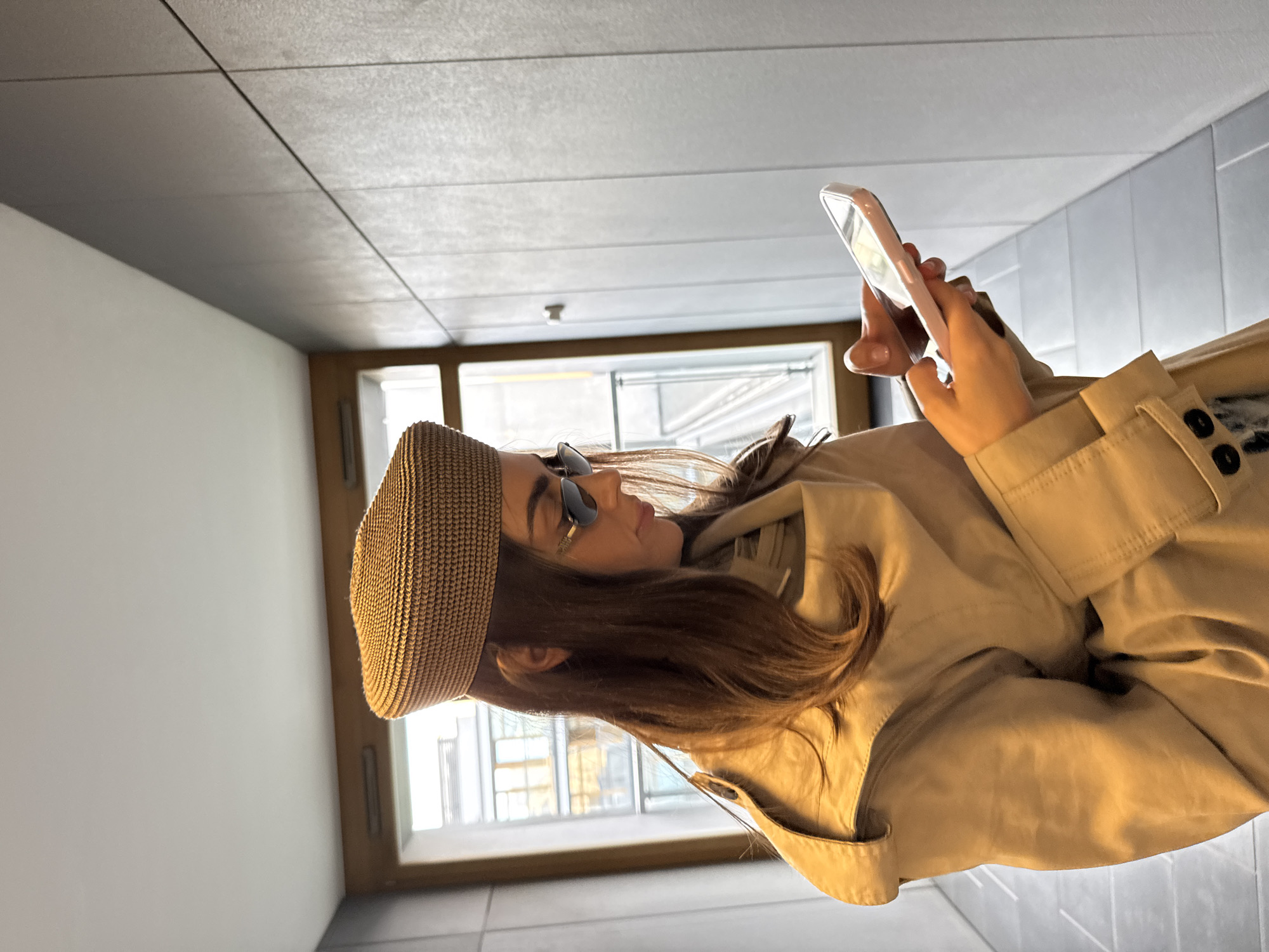 A woman stands in an indoor hallway using a smartphone. She is wearing a light brown trench coat and a woven hat. The hallway has white walls, a gray tiled ceiling, and a window at the end. Outside the window, the cityscape of Unknown, Unknown, is visible.