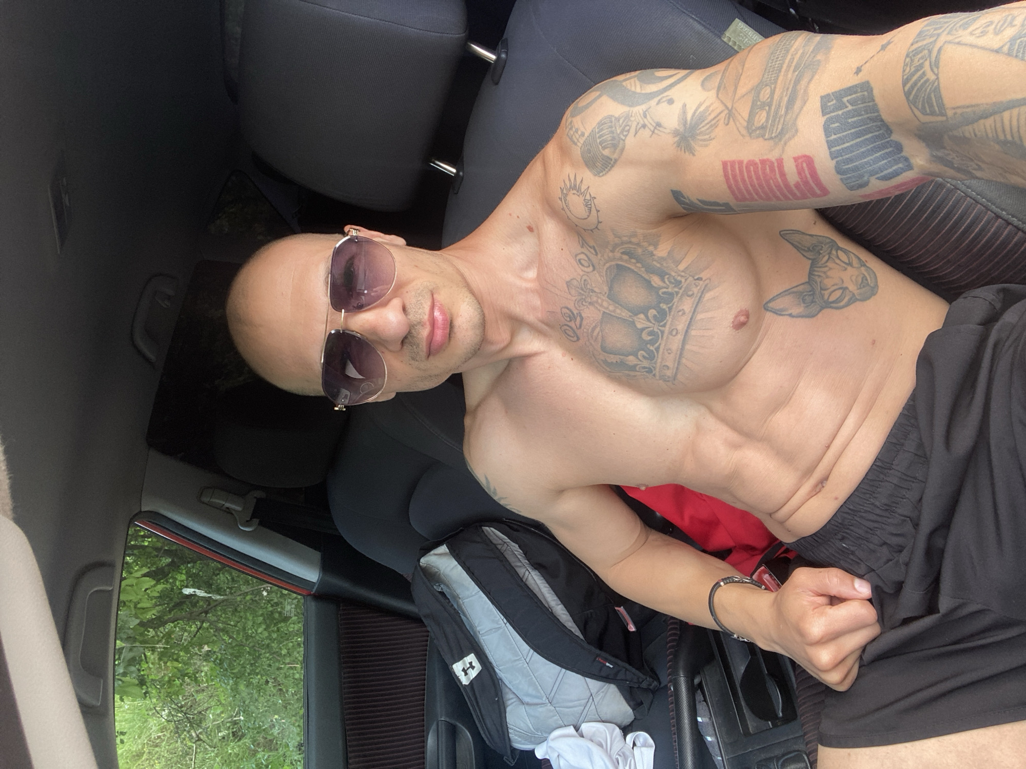 This image depicts a shirtless adult male seated in the driver's side of a parked vehicle, identified by the user as being in the city of Pleven, Bulgaria. The man, with a bald head and wearing mirrored aviator sunglasses, faces forward, capturing a selfie. He exhibits a muscular physique with visible abdominal definition. Numerous tattoos adorn his upper body, including a large crown design with the numbers "02 07" on his chest, a sphinx cat head below his left pectoral, and various designs such as a sun, a microphone, palm trees, a car, and block lettering (partially visible as "WORLD" and "YOURS") on his left arm. A dark bracelet with a metallic accent is visible on his left wrist. He is wearing black athletic shorts.
The car's interior is dark, with dark grey or black seating and a ribbed, possibly dark brown, door panel visible on the left. A grey and black Under Armour backpack rests on the seat next to him, with a white garment on top of it. The center console with cup holders is visible to his lower left. Through the car windows, lush green foliage, consistent with trees and bushes, is visible, indicating an outdoor, natural environment during daylight hours. The passenger seat and rear seating areas are also dark-colored.